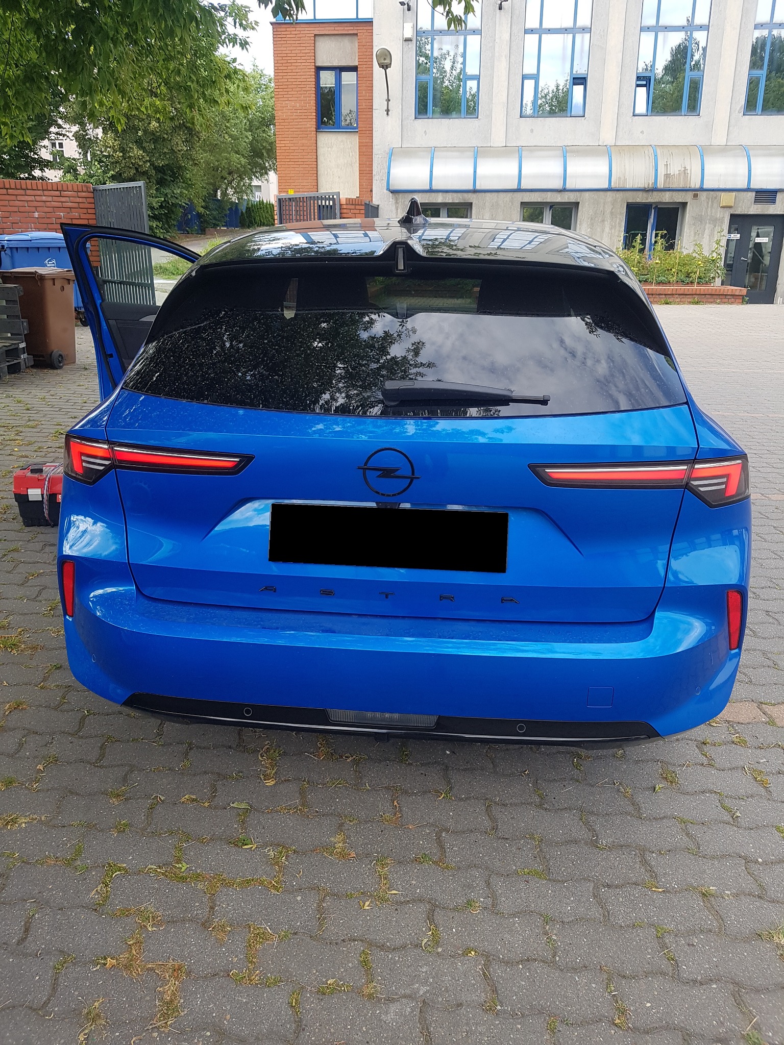 Tył niebieskiego samochodu Opel Astra z otwartymi drzwiami bagażnika, widoczny czerwony pojemnik na narzędzia, na tle budynku z licznymi oknami.