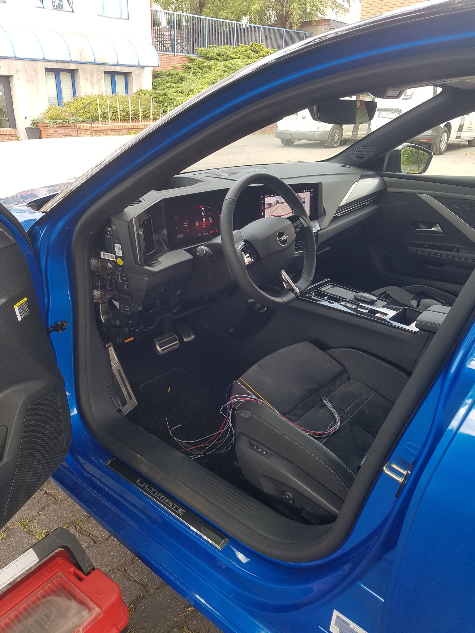 Wnętrze błękitnego samochodu Opel Astra podczas montażu instalacji elektrycznej, widoczne rozpięte przewody na siedzeniu kierowcy i odsłonięta deska rozdzielcza po stronie kierowcy.