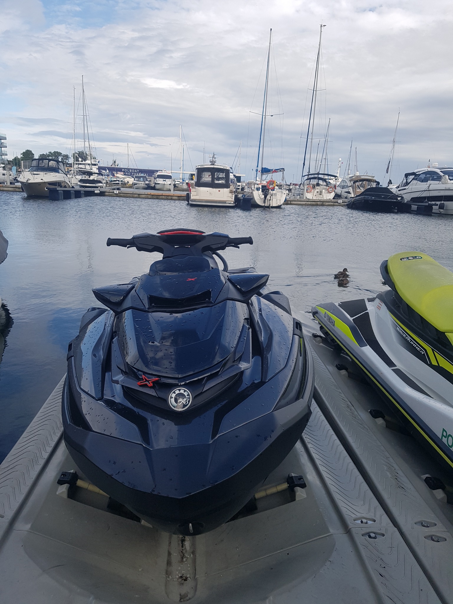 Czarny skuter wodny SeaDoo z kroplami wody na lakierze, zaparkowany w marinie Yacht Park Gdynia, z widokiem na zacumowane jachty i żaglówki.