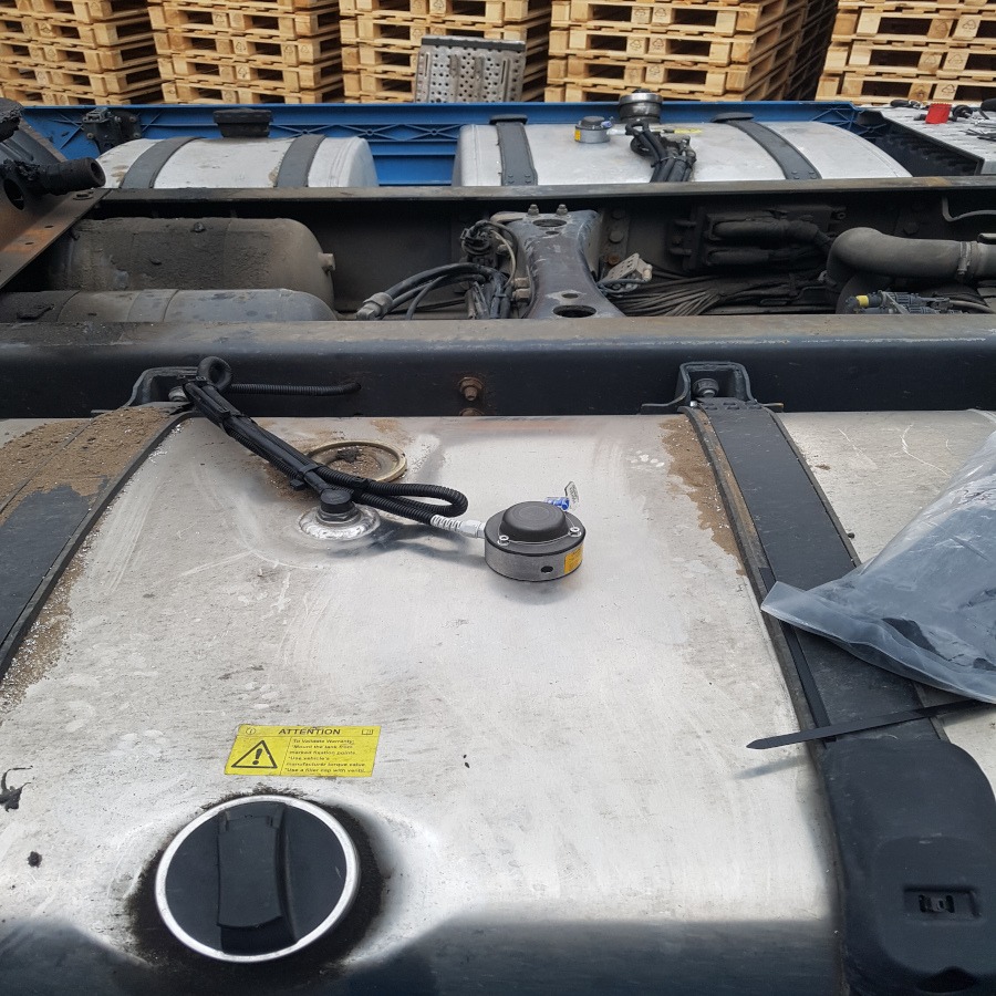 Widok z góry na zamontowany czujnik na zbiorniku paliwa ciężarówki, z widocznymi przewodami i elementami mocującymi, na tle palet.