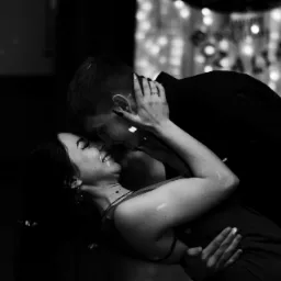 Czarno-białe zdjęcie uśmiechniętej pary, kobieta z kolczykami i pierścionkiem zaręczynowym, mężczyzna w czarnej koszuli, w tle dekoracja z lampek.