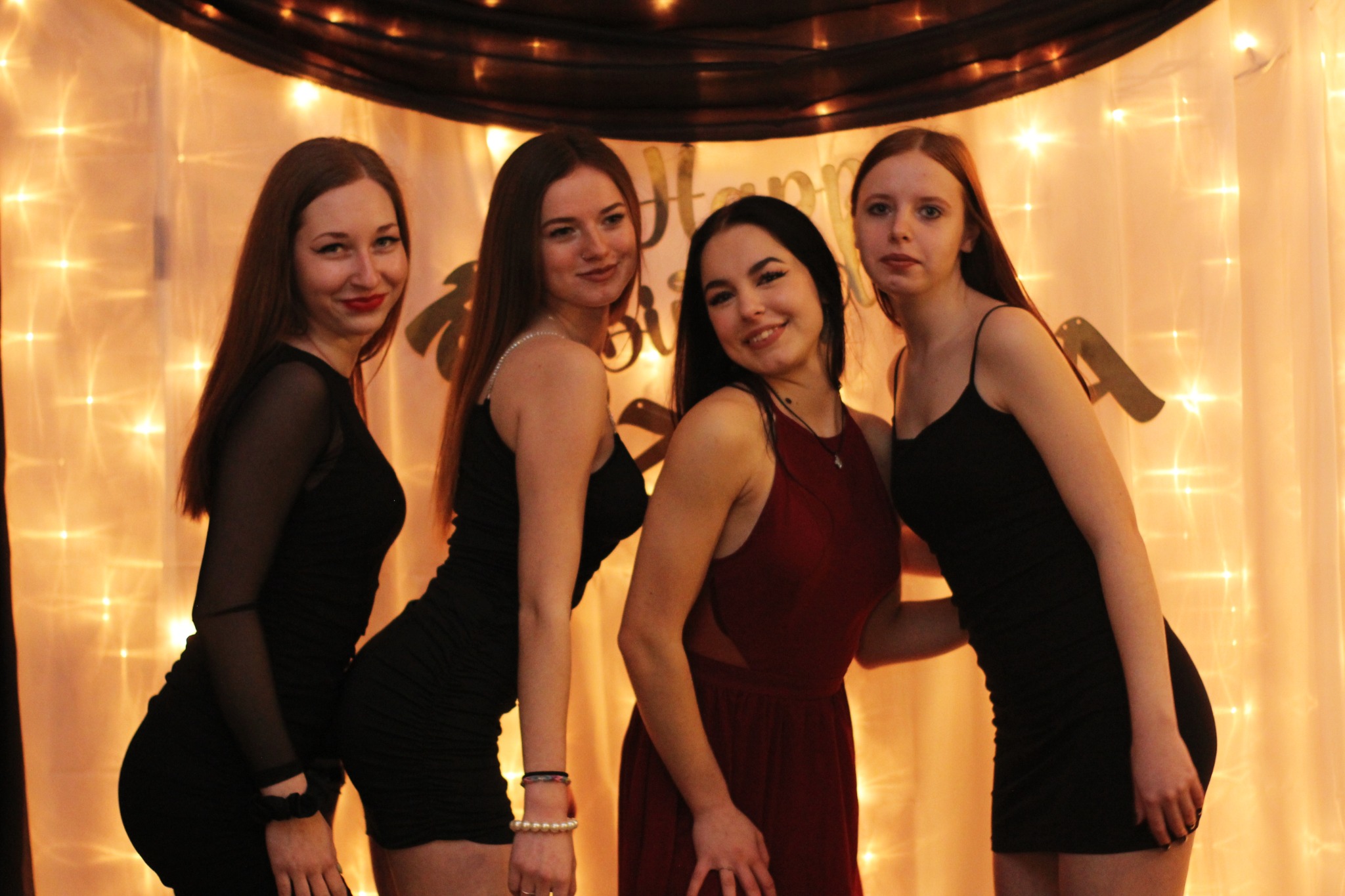 Cztery uśmiechnięte dziewczyny w sukienkach pozują na tle błyszczącej kurtyny z lampek, z dekoracjami urodzinowymi w tle.
