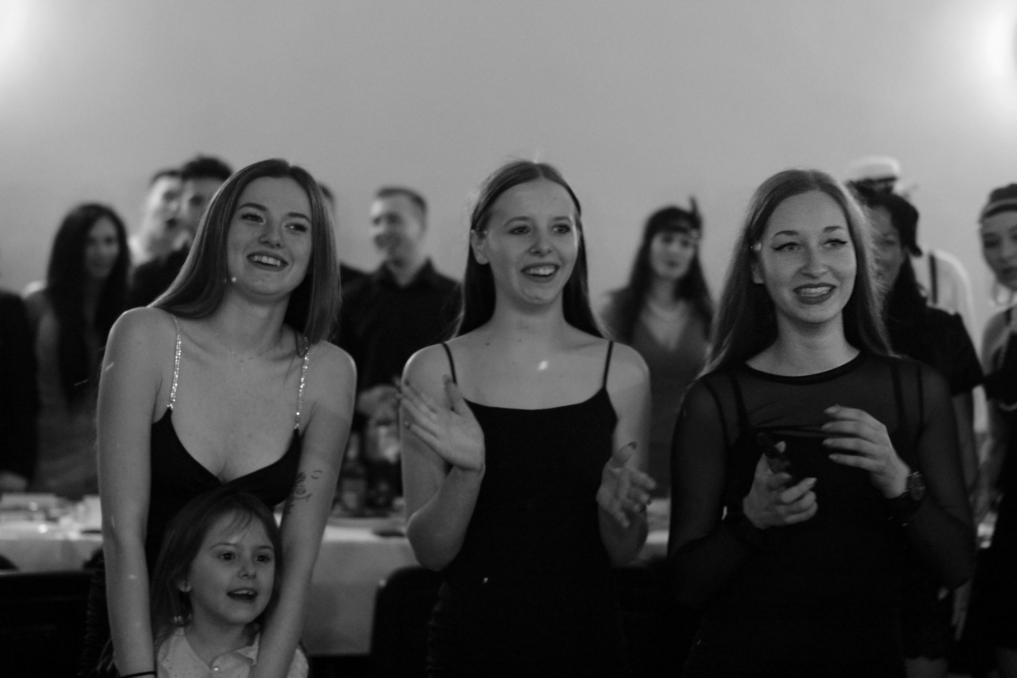 Czarno-białe zdjęcie trzech uśmiechniętych kobiet w czarnych sukienkach, z małą dziewczynką przy nodze jednej z nich, na tle rozmytych postaci i stołów podczas wydarzenia.