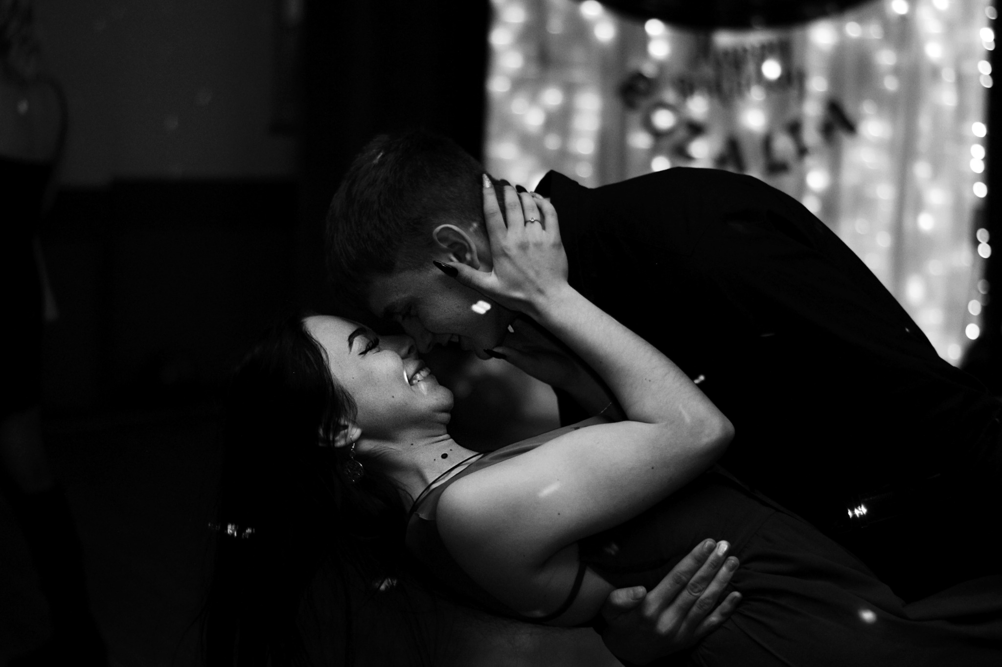 Czarno-białe zdjęcie uśmiechniętej pary, kobieta z kolczykami i pierścionkiem zaręczynowym, mężczyzna w czarnej koszuli, w tle dekoracja z lampek.