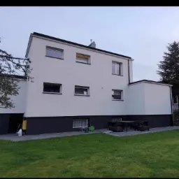 Dwukondygnacyjny budynek z pomalowaną elewacją w kolorze białym i ciemnoszarym cokołem, widoczny z perspektywy trawnika, z oknami o prostokątnym kształcie i ciemnych ramach, w tle drzewa...