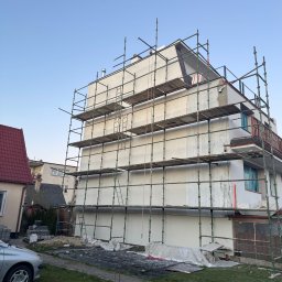Wojtbud - Budynek wielorodzinny w trakcie prac elewacyjnych, otoczony rusztowaniem, z widocznymi śladami świeżo nałożonego tynku i zabezpieczonymi oknami folią, w tle dom z czerwoną dachówką i zaparkowany...