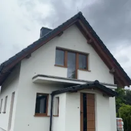 Biały dom po tynkowaniu z brązowymi oknami i drzwiami na tle pochmurnego nieba. Widoczny fragment elewacji z rynną i paletami w tle.