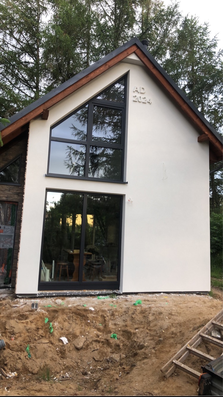 Nowoczesny dom jednorodzinny z jasną elewacją, dużymi oknami w czarnych ramach i datą 'AD 2024' na ścianie szczytowej, widoczny w surowym otoczeniu placu budowy z ziemią i drabiną.