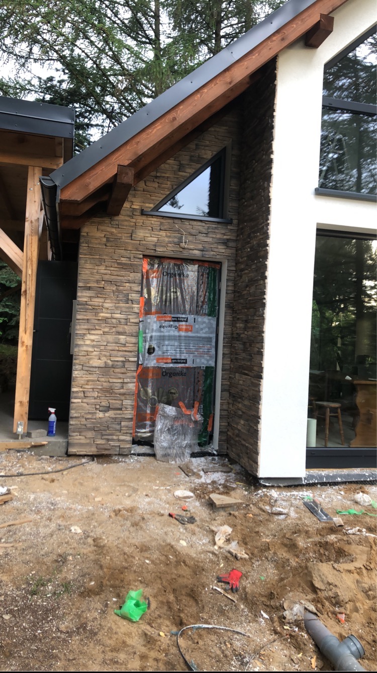 Fragment elewacji domu z wykończeniem imitującym kamień, zabezpieczone drzwi wejściowe folią ochronną, widoczne narzędzia i materiały budowlane na ziemi.