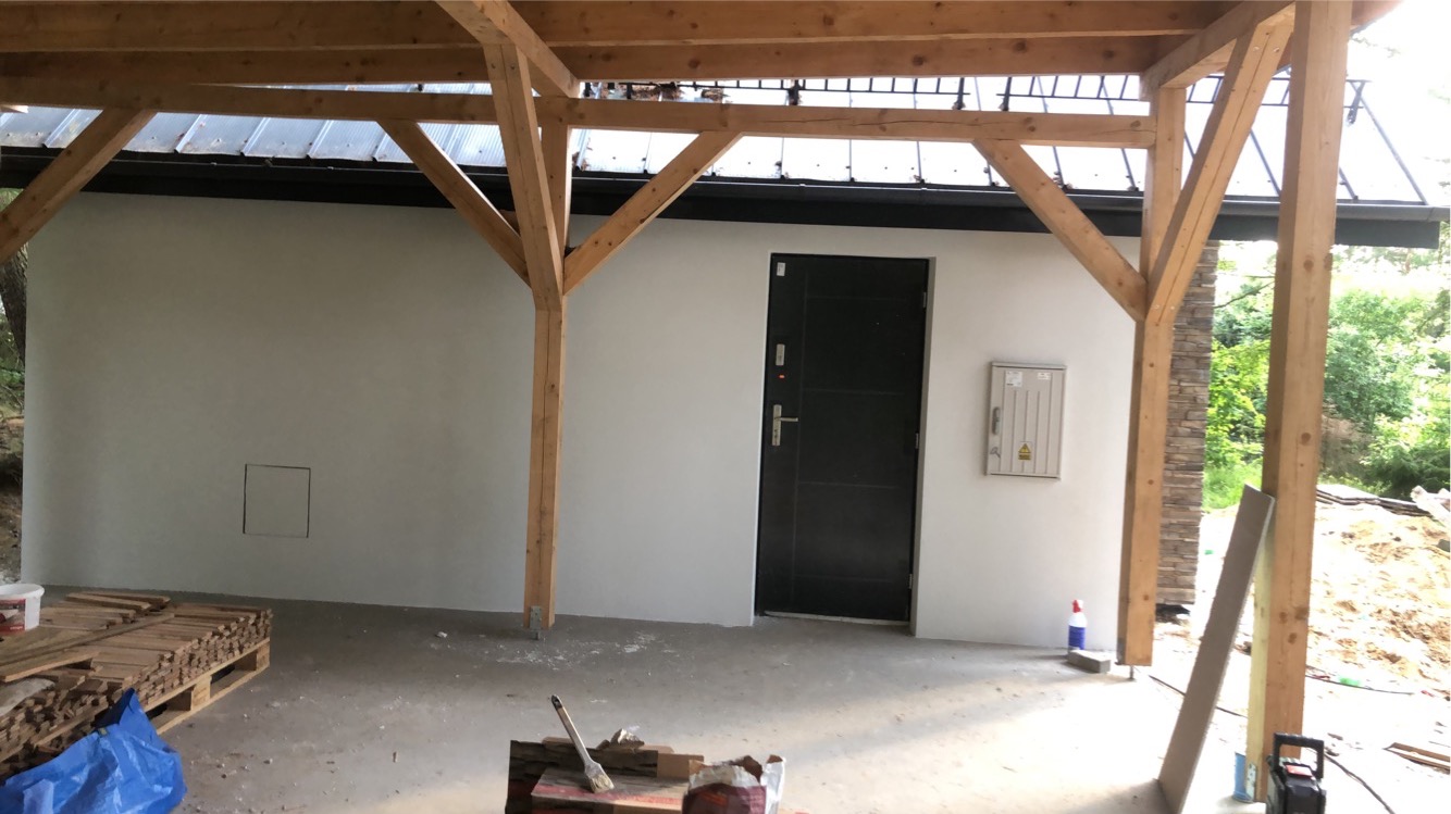 Świeżo otynkowana ściana zewnętrzna budynku z drewnianą konstrukcją zadaszenia, czarne drzwi wejściowe i skrzynka elektryczna na elewacji, widoczne materiały budowlane i narzędzia malarskie.