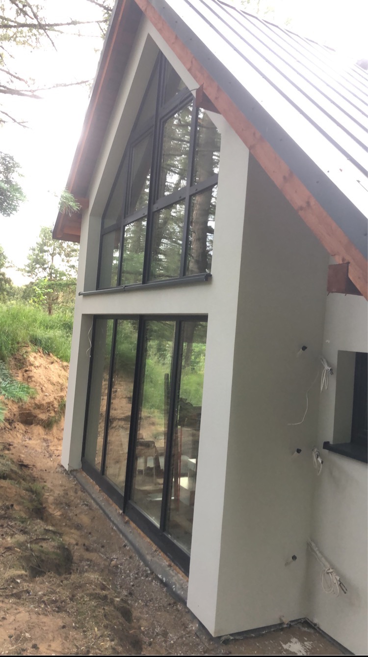 Nowoczesny dom z dużymi przeszkleniami, świeżo otynkowaną elewacją w odcieniach szarości, widok z boku na tle zieleni i drzew, z widocznymi przewodami elektrycznymi na zewnętrznej ścianie.