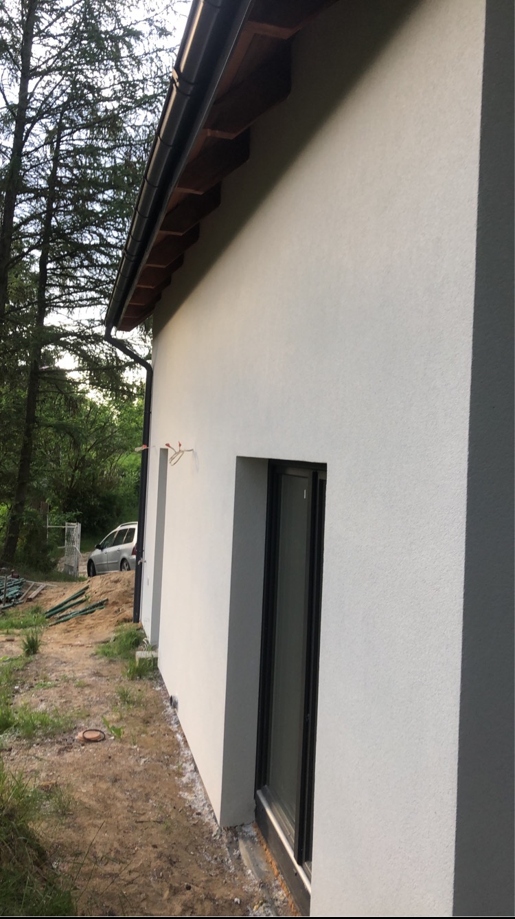 Świeżo otynkowana, jasnoszara elewacja domu jednorodzinnego z czarną stolarką okienną i orynnowaniem, widoczne wystające przewody elektryczne, teren wokół domu jeszcze niezagospodarowany.