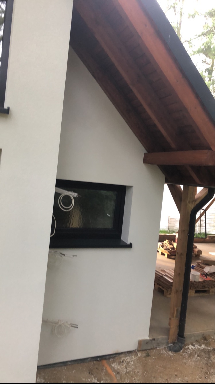 Fragment budynku z jasną elewacją, oknem w czarnej ramie i drewnianym zadaszeniem, widoczne wystające przewody i przygotowany materiał budowlany.