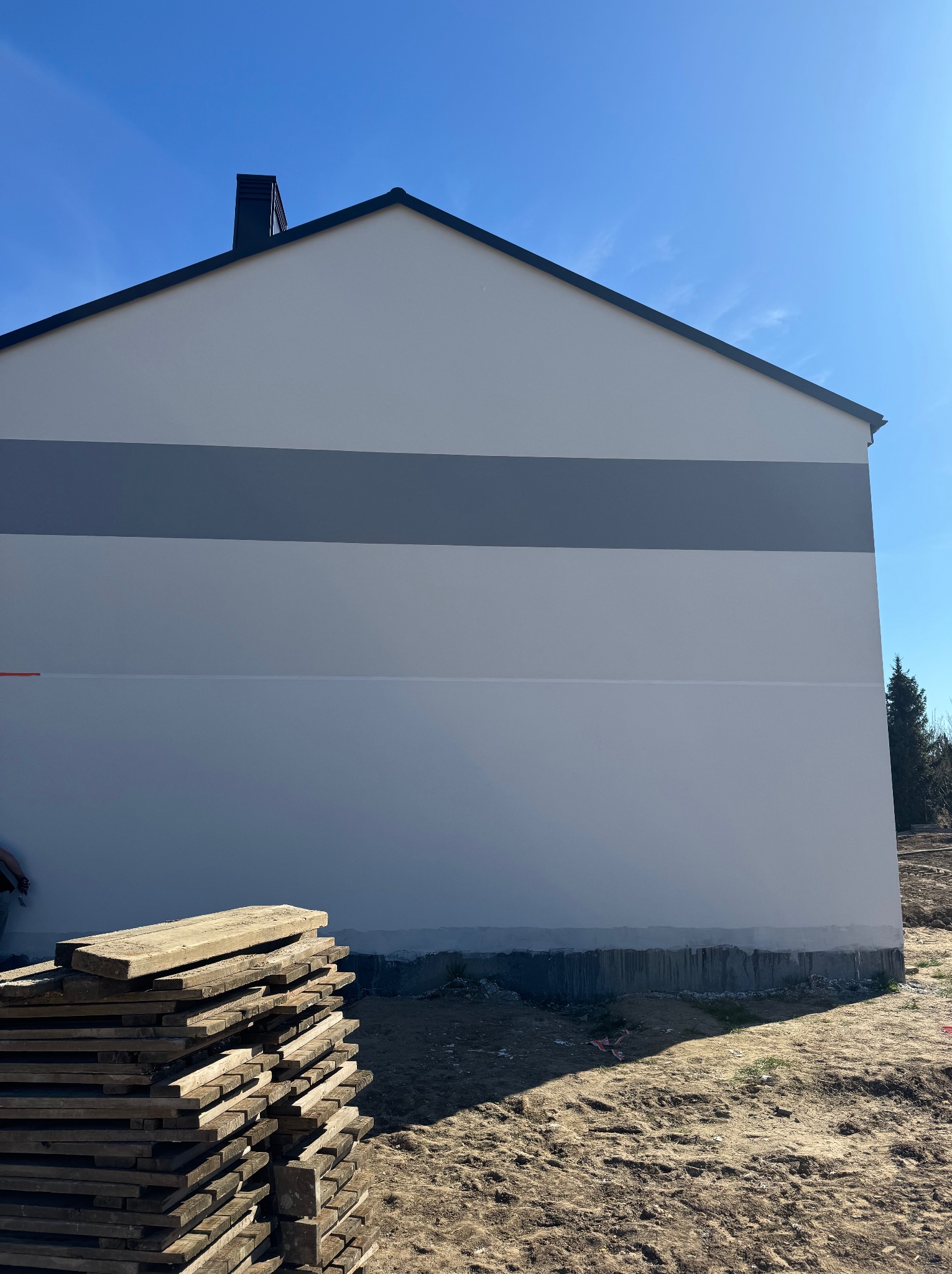 Świeżo pomalowana elewacja budynku z szarym pasem dekoracyjnym, widoczny komin i stos desek na pierwszym planie, błękitne niebo w tle.