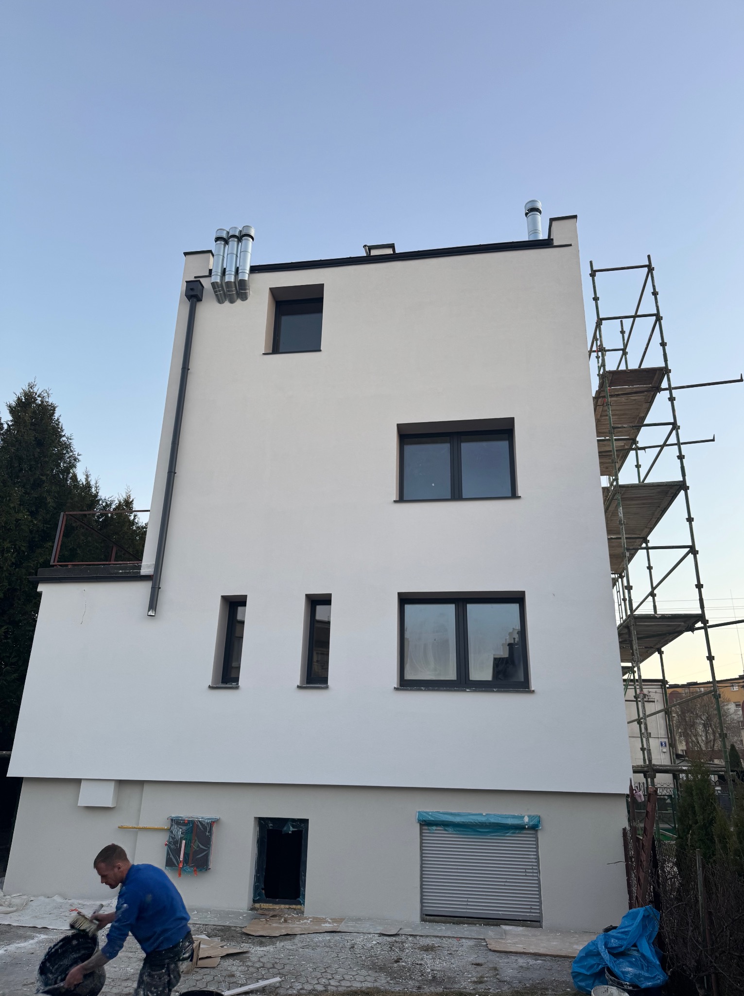 Pracownik w niebieskiej bluzie przygotowuje zaprawę przed budynkiem z nową, jasną elewacją, widoczne rusztowania i trzy kominy na dachu.