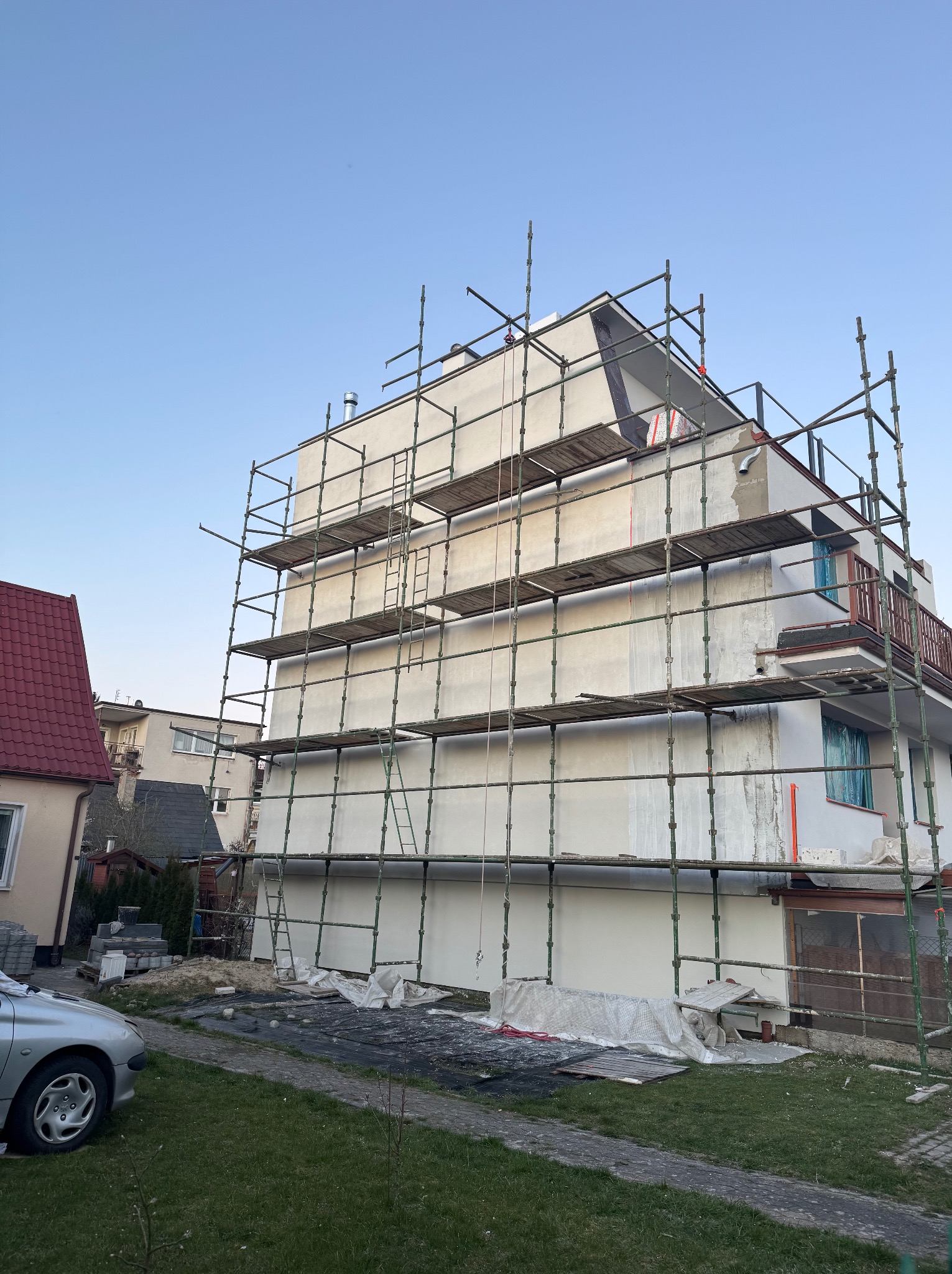 Budynek wielorodzinny w trakcie prac elewacyjnych, otoczony rusztowaniem, z widocznymi śladami świeżo nałożonego tynku i zabezpieczonymi oknami folią, w tle dom z czerwoną dachówką i zaparkowany...
