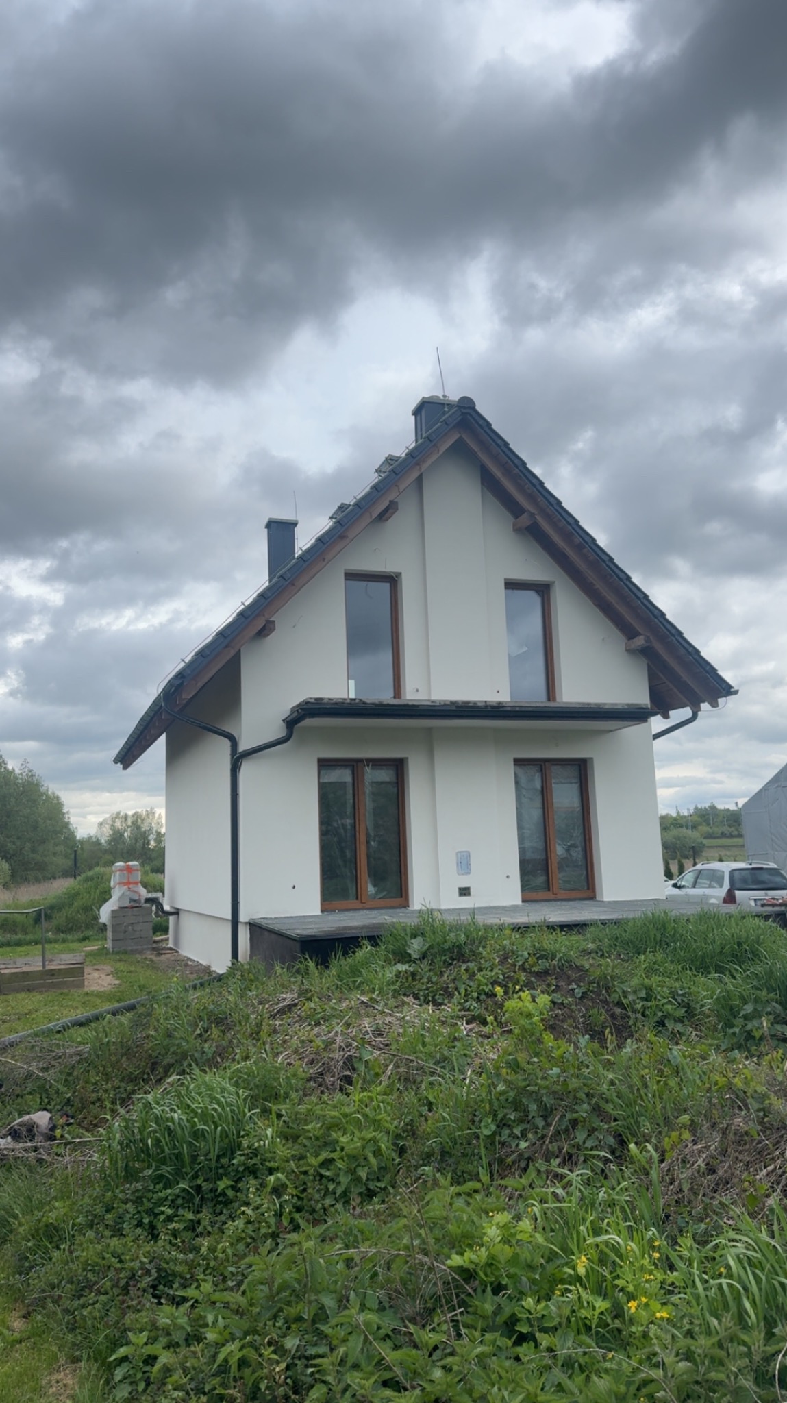 Biały dom jednorodzinny z brązowymi oknami i ciemnym dachem na tle pochmurnego nieba, otoczony zielenią. Widoczne elementy wykończeniowe elewacji.