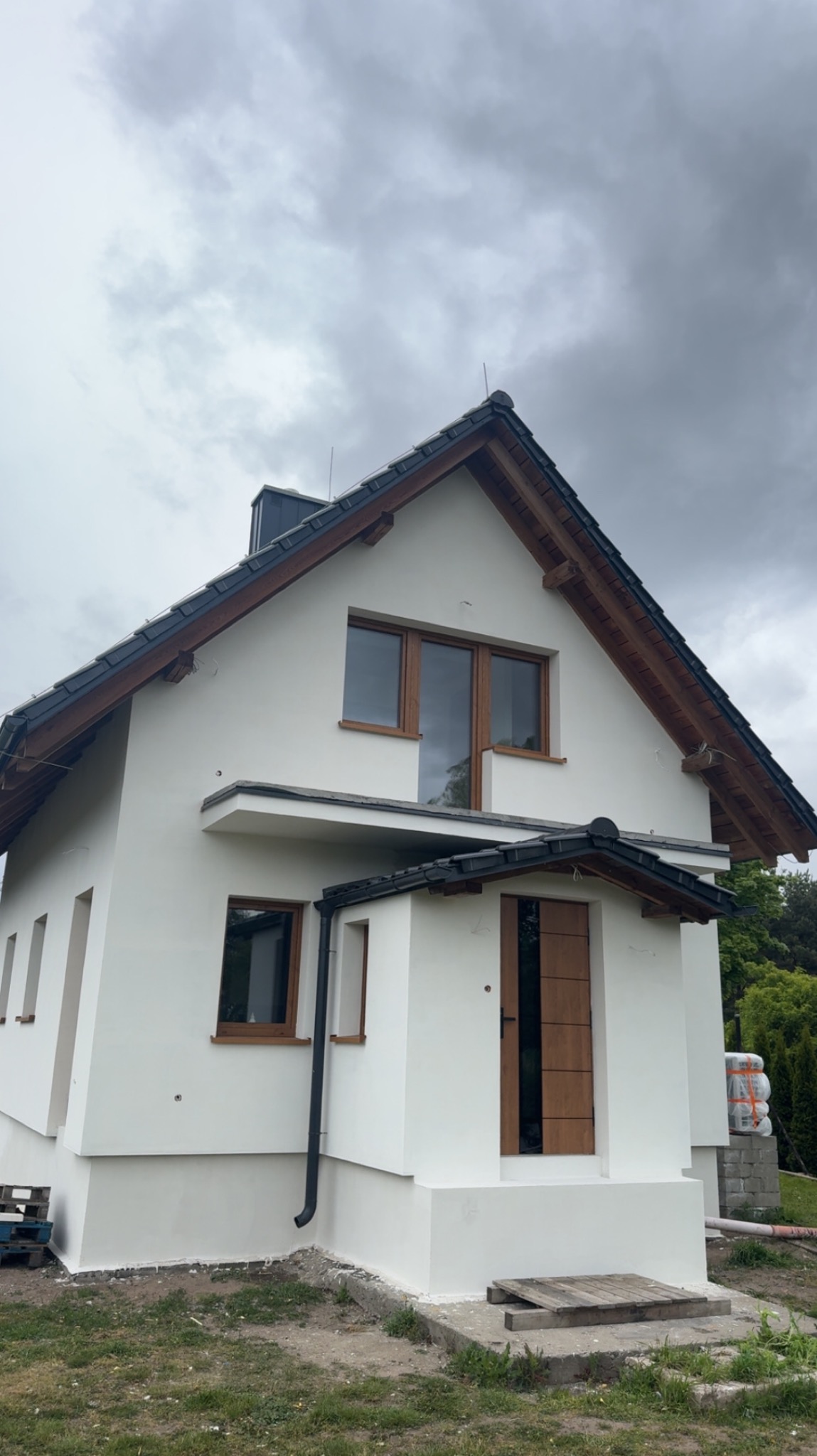 Biały dom po tynkowaniu z brązowymi oknami i drzwiami na tle pochmurnego nieba. Widoczny fragment elewacji z rynną i paletami w tle.