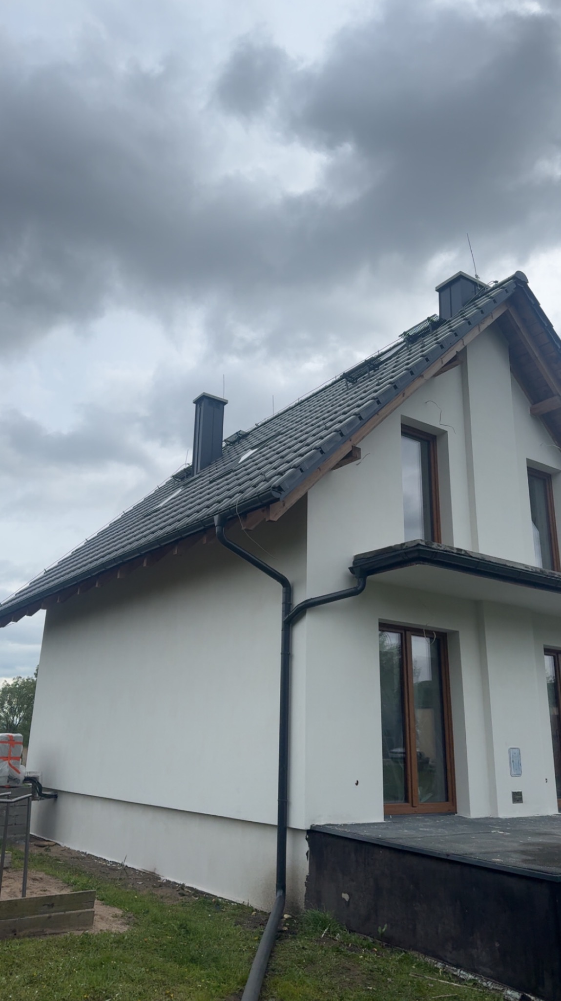 Elewacja domu jednorodzinnego po tynkowaniu, widok z boku na tle pochmurnego nieba. Widoczne okna z brązowymi ramami i czarne rynny.