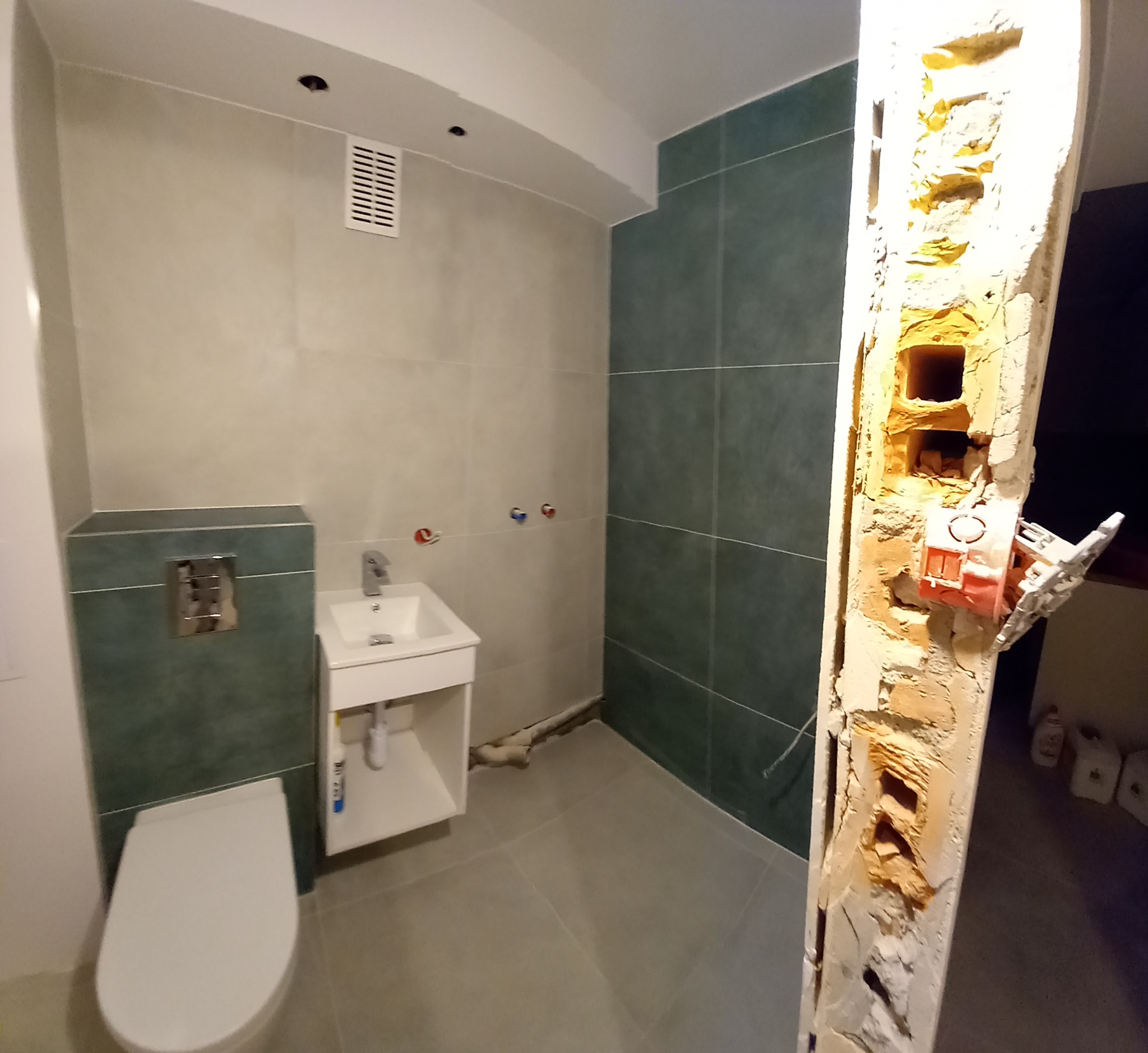 Wnętrze łazienki w trakcie prac, z widoczną toaletą, umywalką, szarą i zieloną glazurą oraz fragmentem ściany z odsłoniętą instalacją elektryczną.