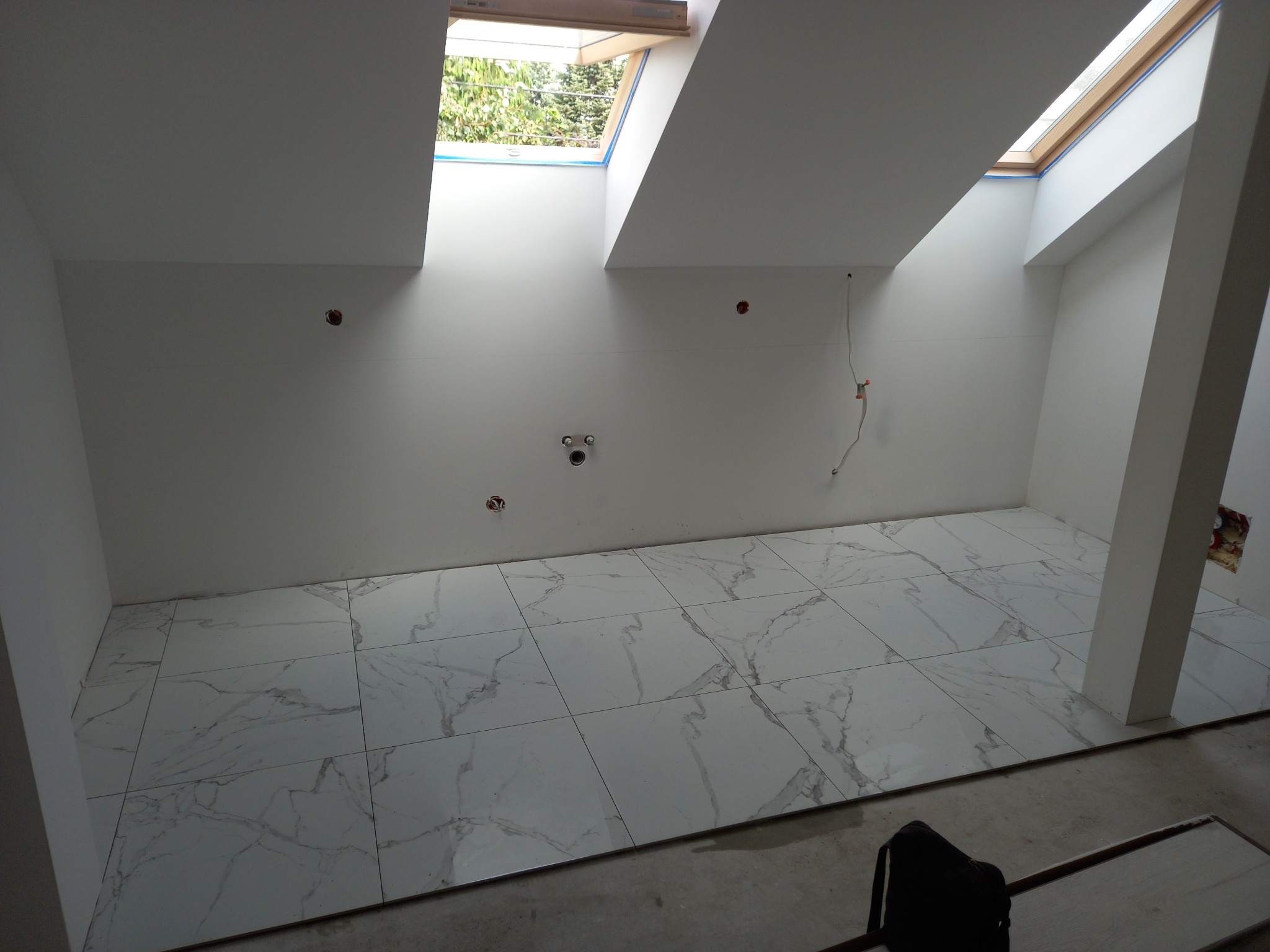 Poddasze z ułożoną białą marmurkową podłogą z płytek, widoczne okna dachowe i przygotowane punkty instalacji elektrycznej i hydraulicznej na ścianach.