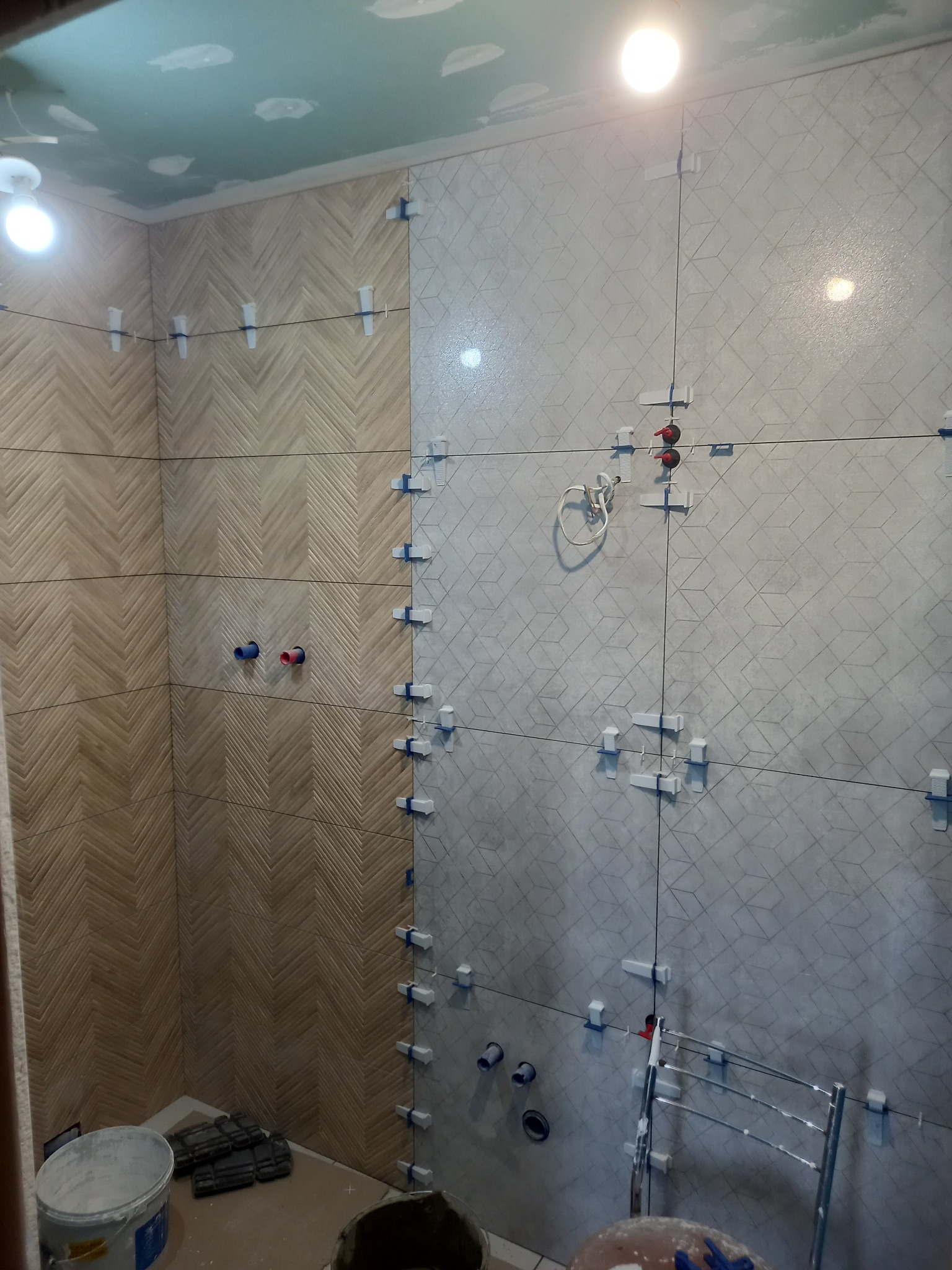 Łazienka w trakcie kładzenia płytek: wzór jodełki z drewna oraz geometryczny wzór 3D, widoczne krzyżyki dystansowe i poziomica.