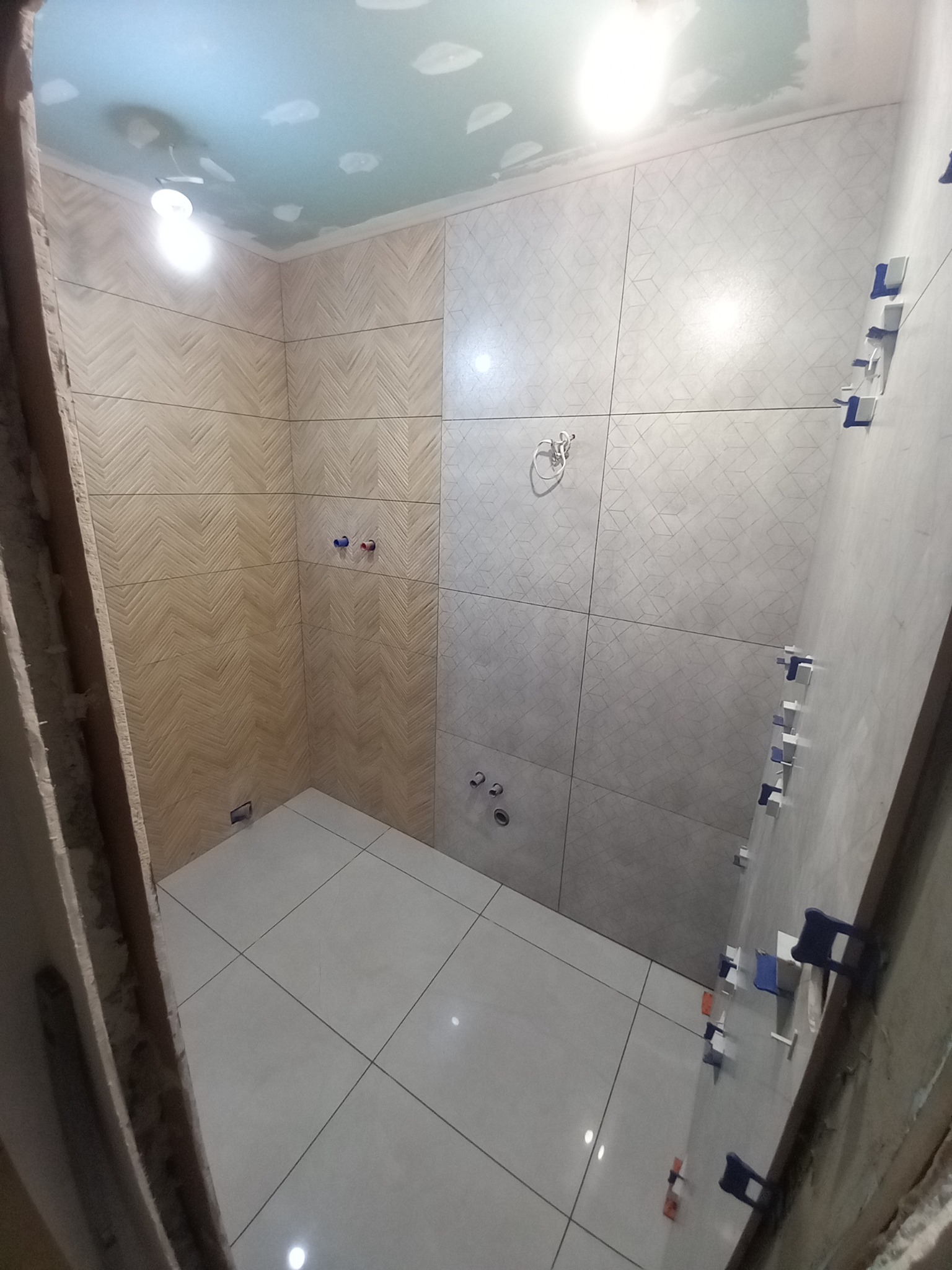 Łazienka w trakcie wykańczania: dwie ściany z ułożonymi płytkami - jedna z wzorem jodełki, druga z geometrycznym, jasne płytki podłogowe, widoczne krzyżyki dystansowe na jednej ze ścian, sufit...