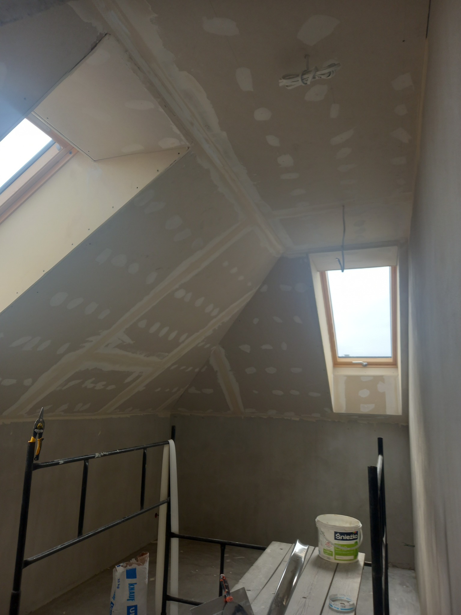 Poddasze w trakcie prac wykończeniowych: ściany i sufit pokryte płytami gipsowo-kartonowymi, częściowo zaszpachlowane, widoczne dwa okna dachowe, rusztowanie, wiadro z farbą Śnieżka i narzędzia.