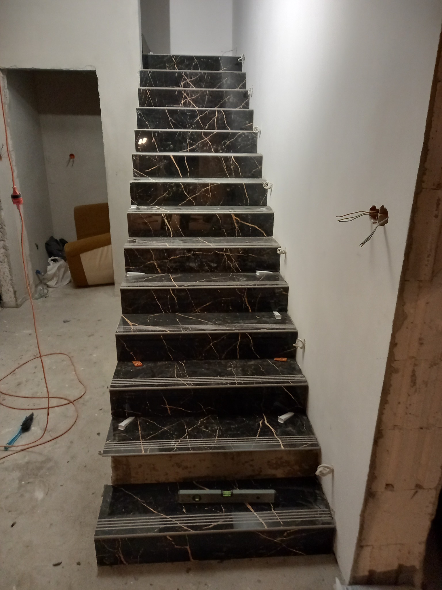 Schody wyłożone czarnymi płytkami imitującymi marmur z wyraźnym złotym użyleniem, w trakcie prac wykończeniowych, widoczne poziomica i fragment kabla elektrycznego wystającego ze ściany.