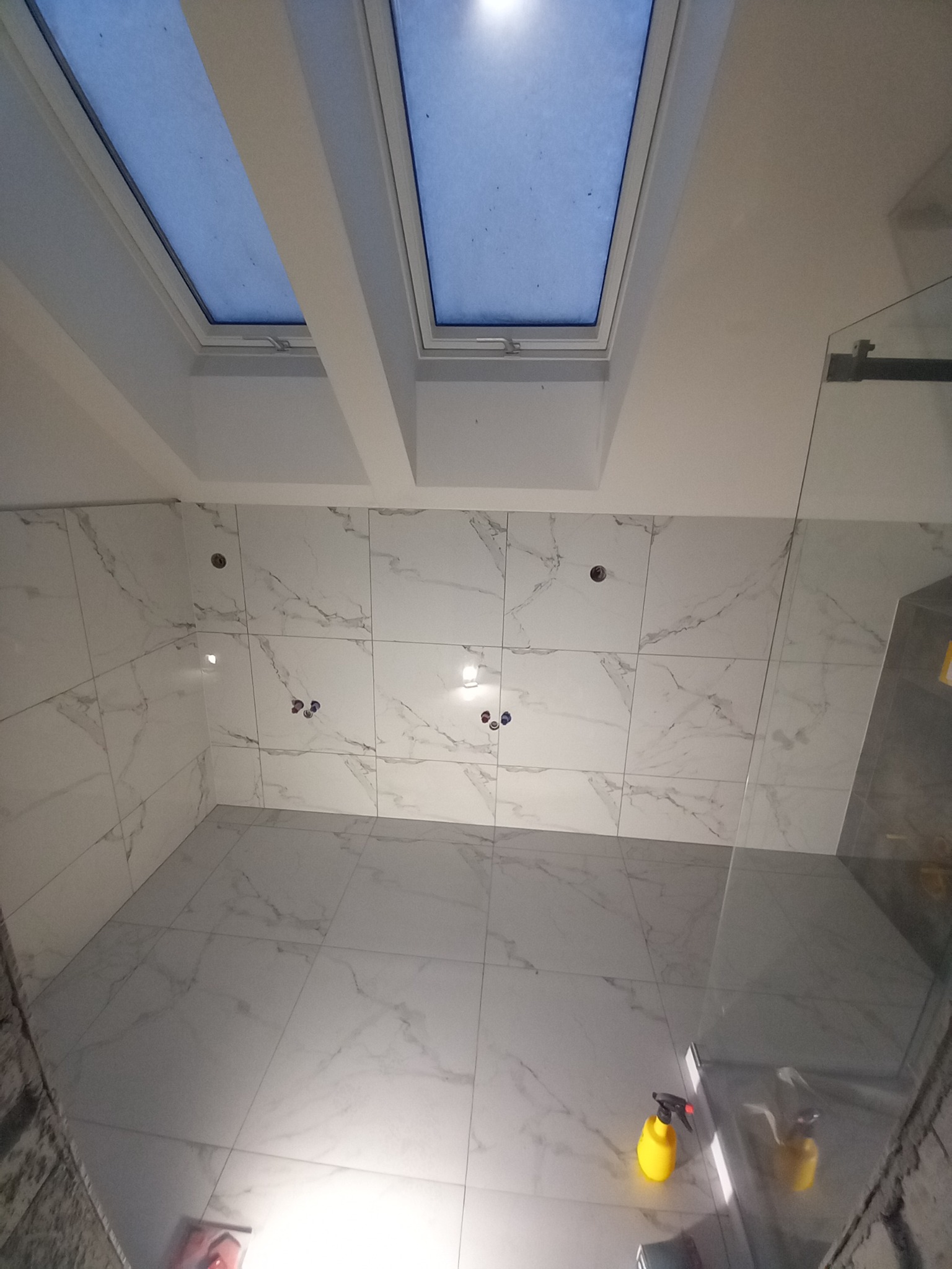 Wykończona łazienka w Łodzi z białymi płytkami imitującymi marmur na ścianach i podłodze, widoczne dwa okna dachowe i częściowo szklana kabina prysznicowa, przygotowana do użytku.