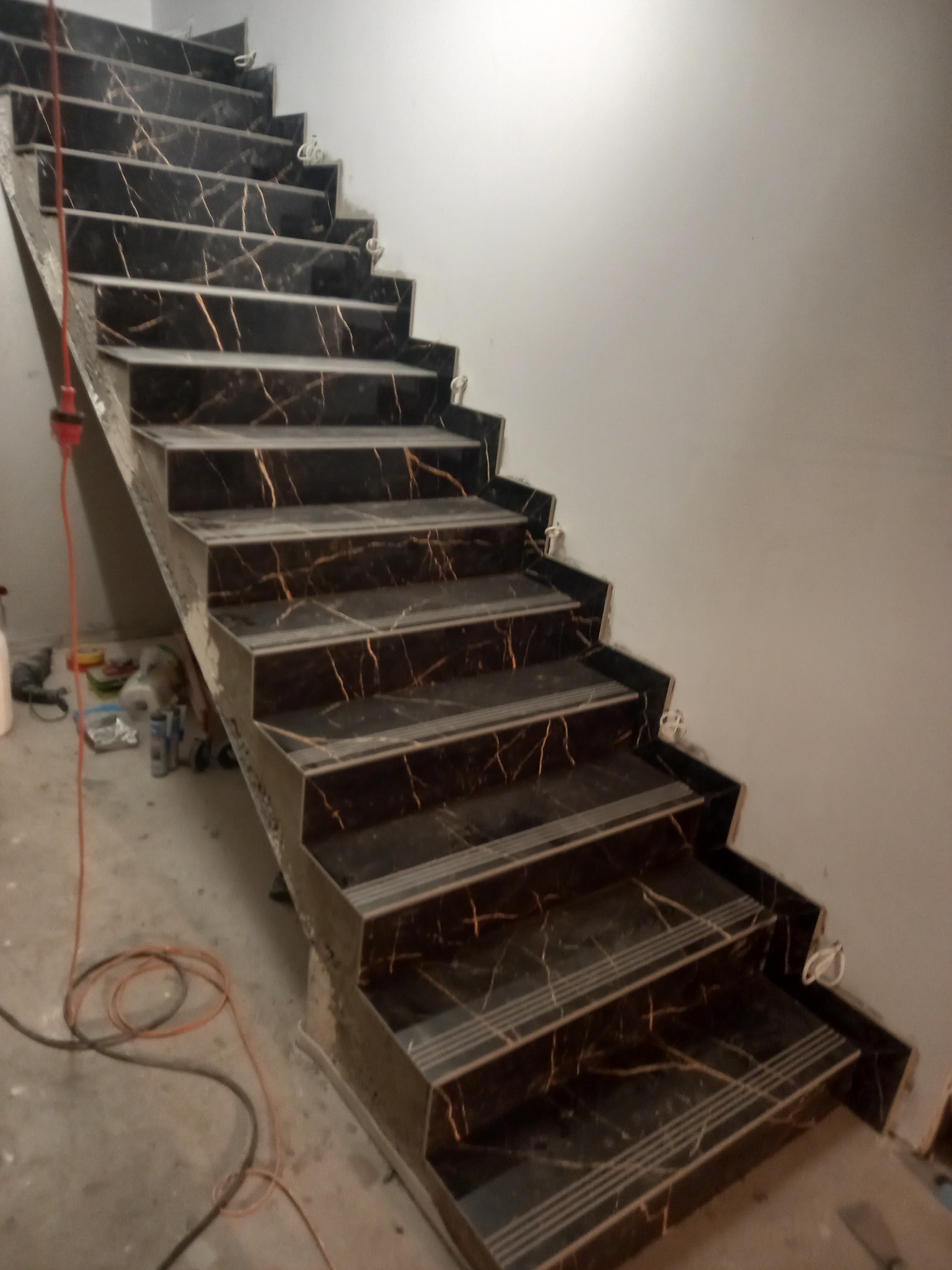 Schody wyłożone czarnymi płytkami imitującymi marmur ze złotymi żyłami, widoczne narzędzia i przewody na posadzce, ściana w tle w odcieniach szarości.