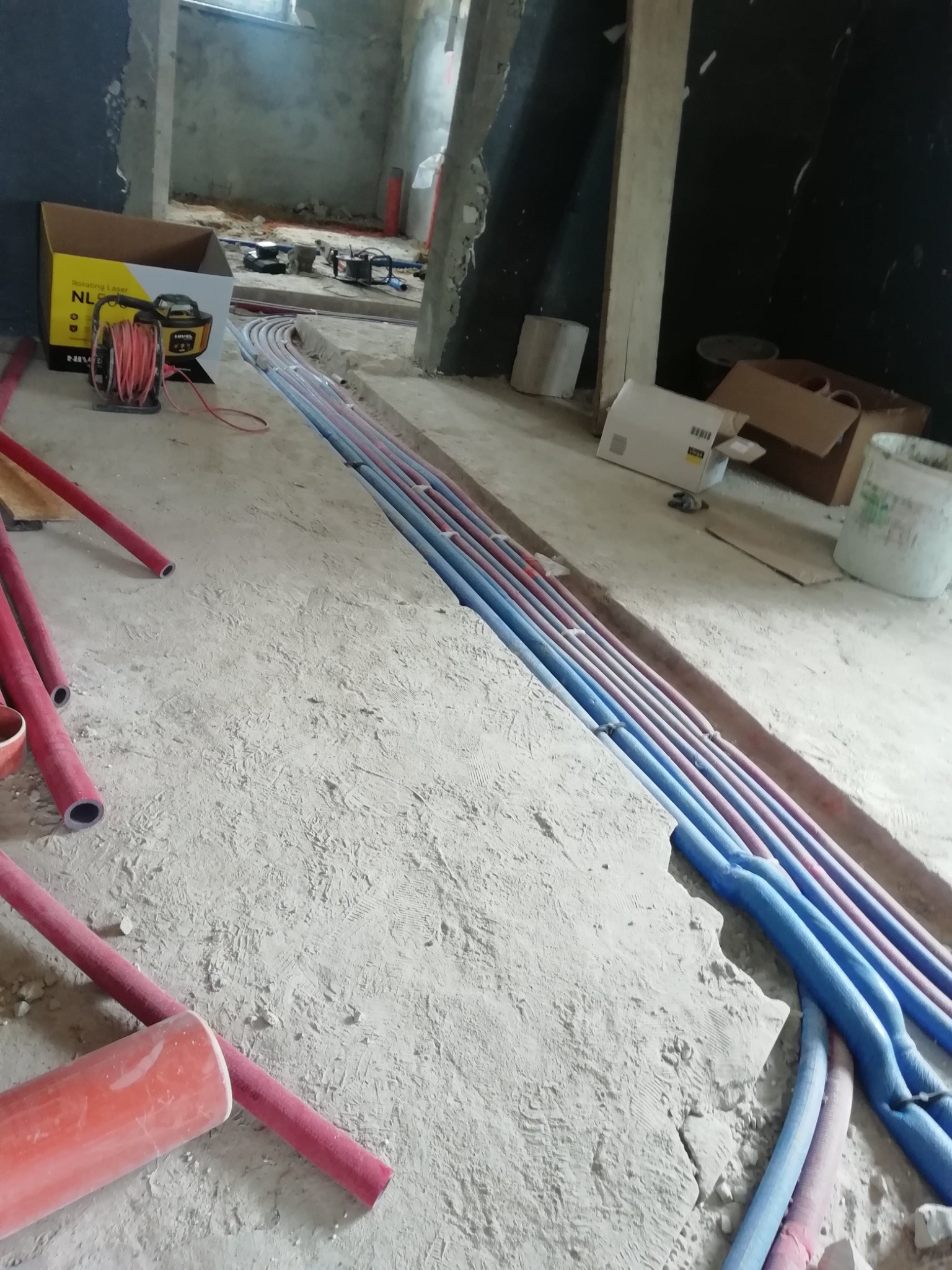 Wykop w surowym betonie z ułożonymi rzędami niebieskich i czerwonych rur instalacji wodnej i grzewczej, widoczny laser krzyżowy i zwój czerwonego przewodu w tle.