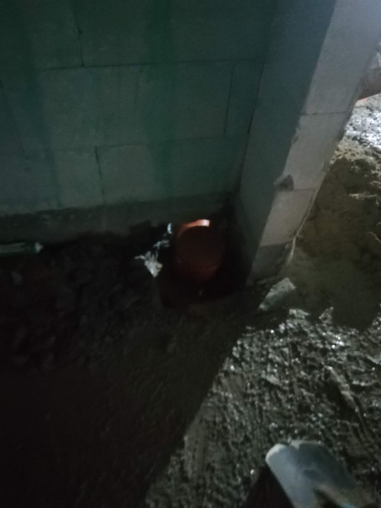 Pomarańczowa rura kanalizacyjna w wykopie przy betonowej ścianie fundamentowej, widoczne mokre podłoże i fragment szpadla.