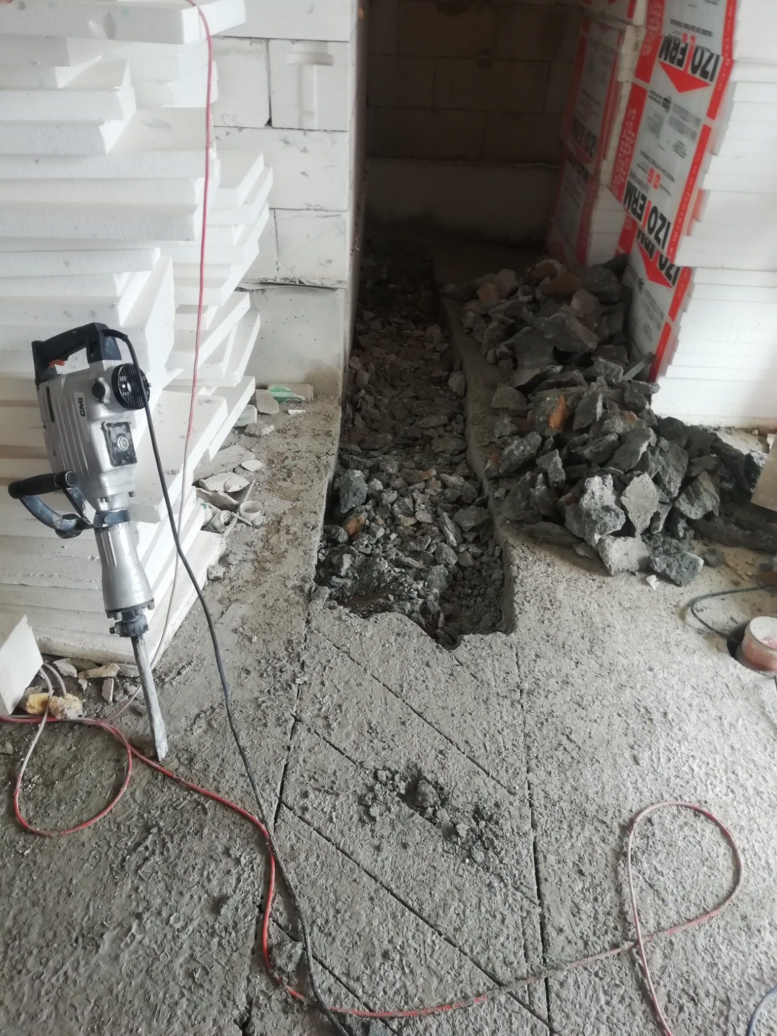 Elektryczny młot udarowy stoi obok wyżłobionego w betonie koryta, z gruzem i fragmentami betonu wewnątrz, w tle białe płyty styropianowe i izolacja IZO ERM.