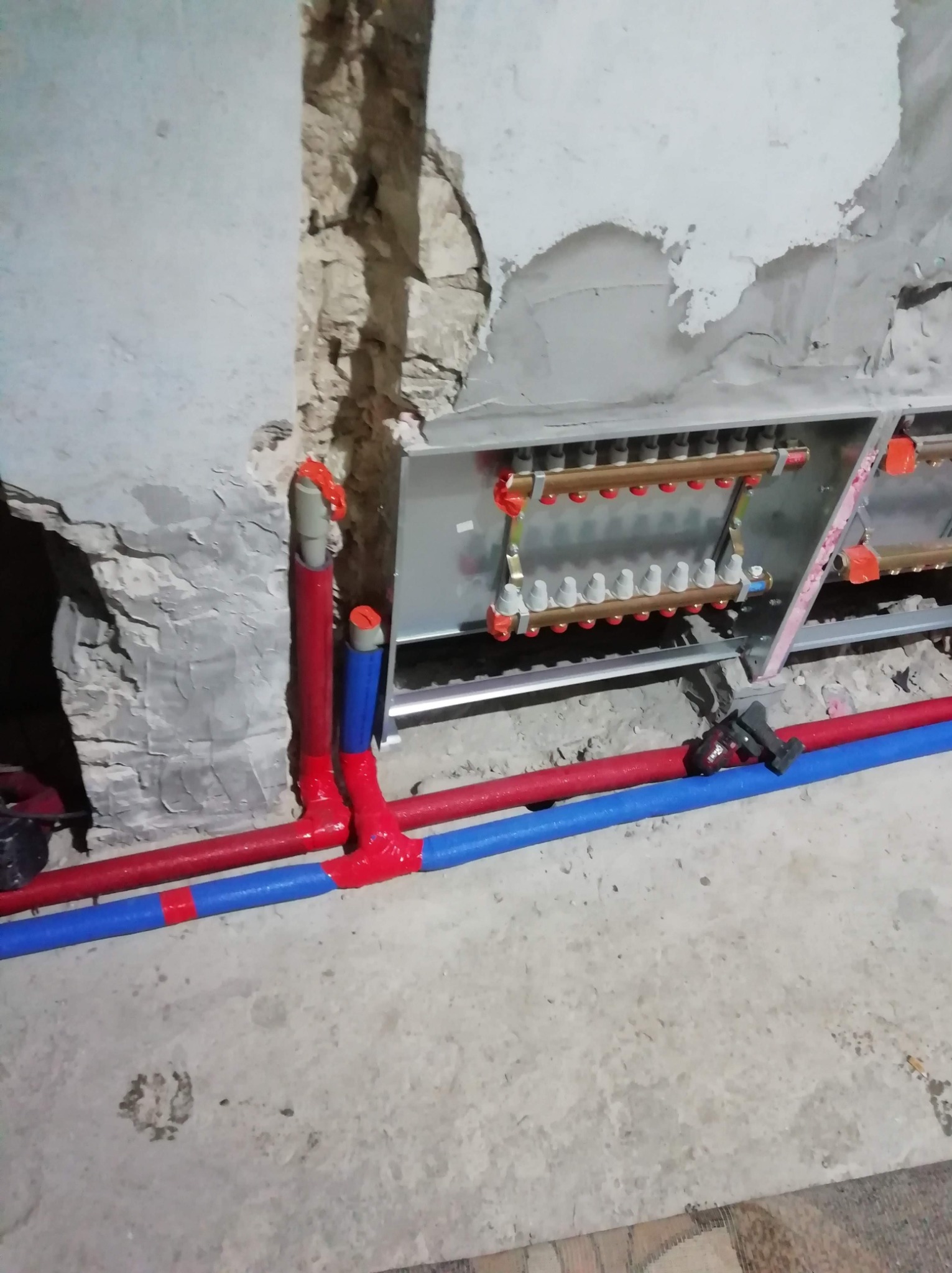Rozdzielacz instalacji centralnego ogrzewania, rury w kolorze czerwonym i niebieskim, fragment ściany w stanie surowym, widoczne ślady po kuciu.