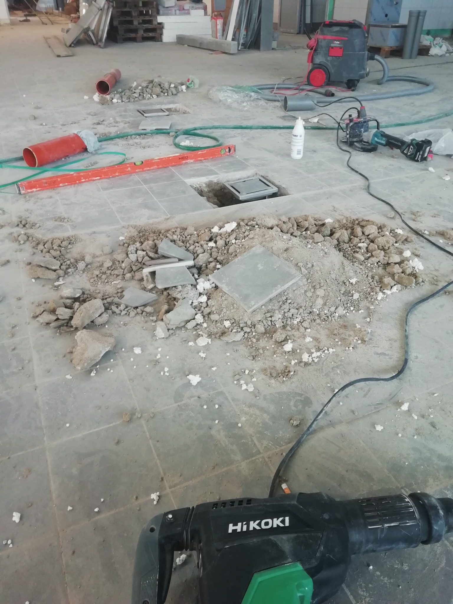 Demontaż podłogi z płytek: gruz, rury, poziomica, odkurzacz przemysłowy, wiertarka udarowa HiKOKI i wycięty otwór w posadzce.