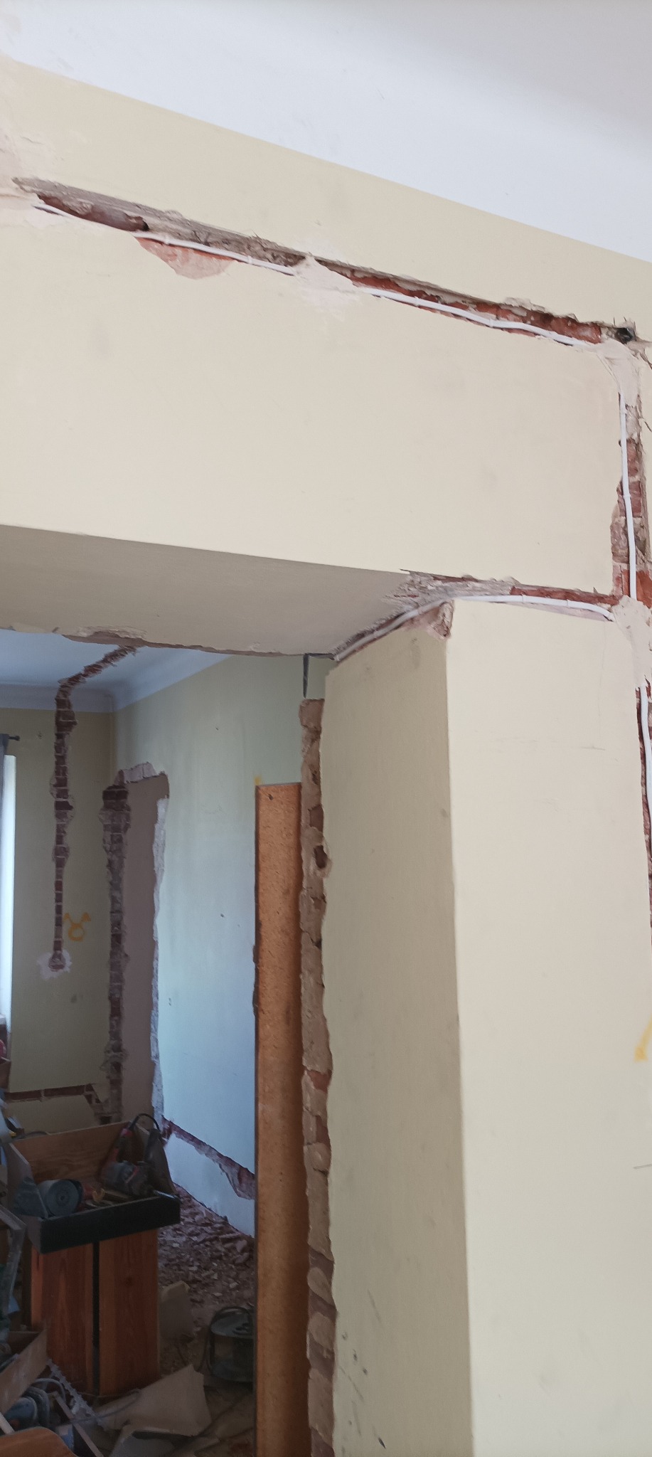 Ściana po kuciu bruzd pod instalacje elektryczne, widoczne przewody w wyżłobieniach, fragmenty cegieł i resztki zaprawy. W tle widoczny bałagan budowlany.