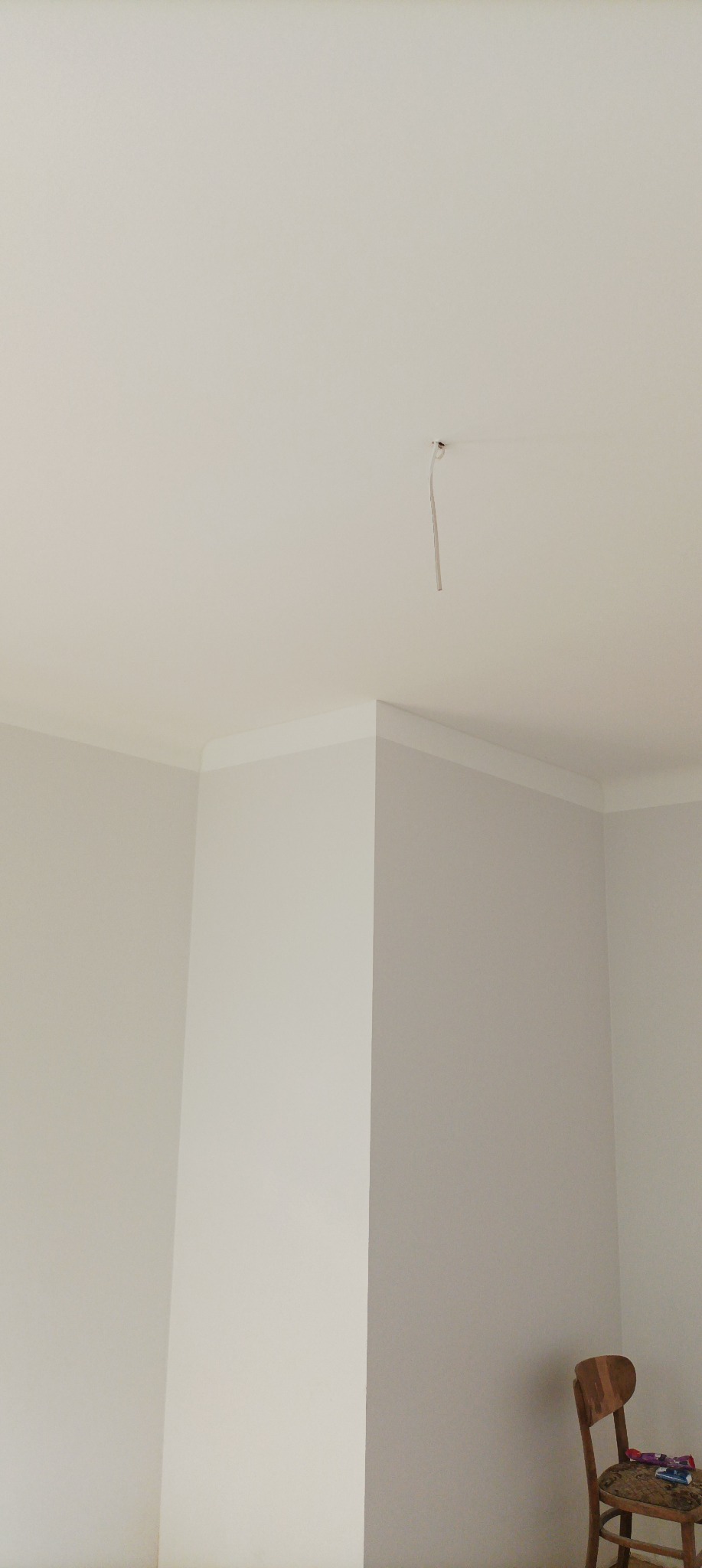 Wnętrze pokoju w trakcie remontu: biały sufit z wystającym kablem elektrycznym, świeżo pomalowane ściany z dekoracyjną listwą przysufitową, krzesło w rogu.