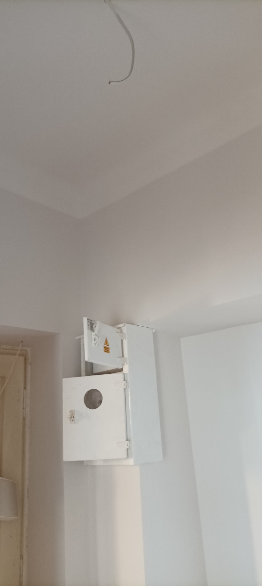 Biała skrzynka elektryczna z otwartymi drzwiczkami, zamontowana na ścianie w rogu pomieszczenia, widoczny wystający kabel z sufitu.