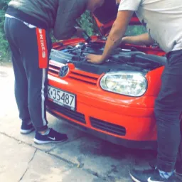 Dwóch mężczyzn pochylonych nad otwartą maską czerwonego Volkswagena Golfa, sprawdzających silnik na zewnątrz warsztatu, widoczna tablica rejestracyjna K-35487