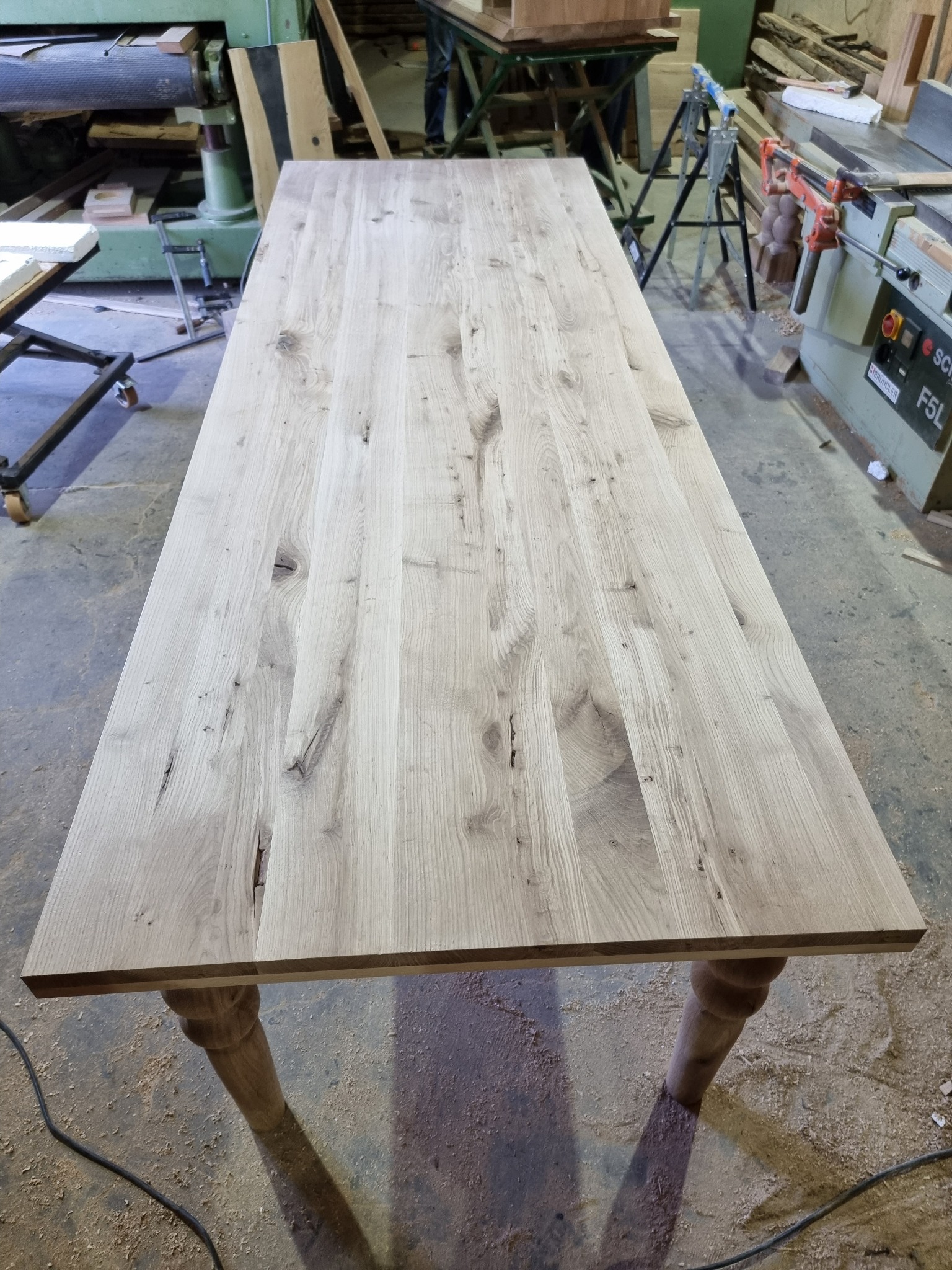 Blat stołu z jasnego drewna z sękami, wykonany w warsztacie w Modliborzycach, widoczne toczone nogi i maszyny stolarskie w tle.