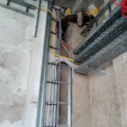 Kable elektryczne prowadzone w metalowych korytkach i mocowane do ściany, widoczna izolacja w ścianie, surowy stan wykończenia wnętrza.