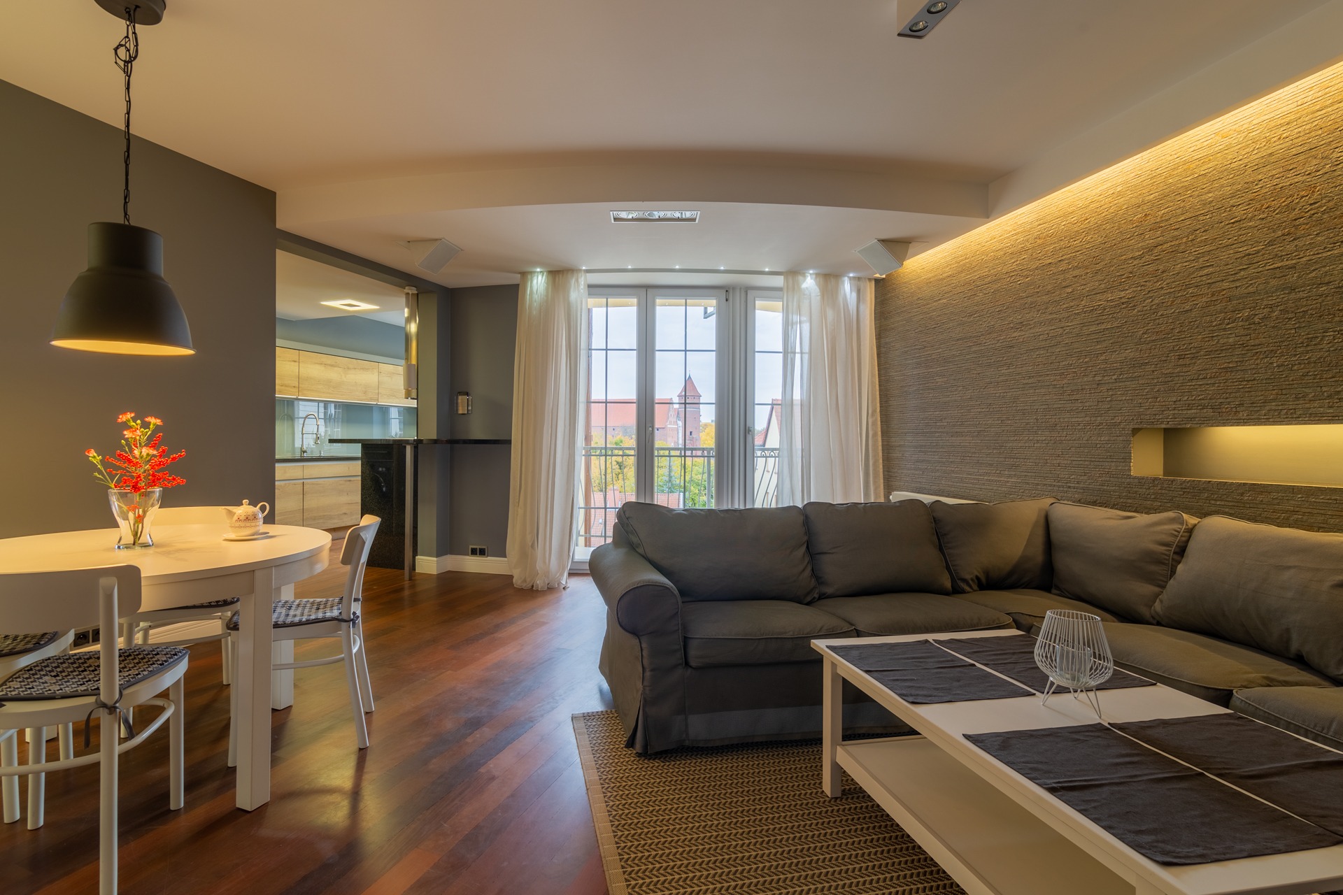 Jasne wnętrze mieszkania z drewnianą podłogą, szarą sofą narożną, białym stolikiem kawowym i okrągłym stołem jadalnym z kwiatami, widok z okna na ceglaną wieżę.