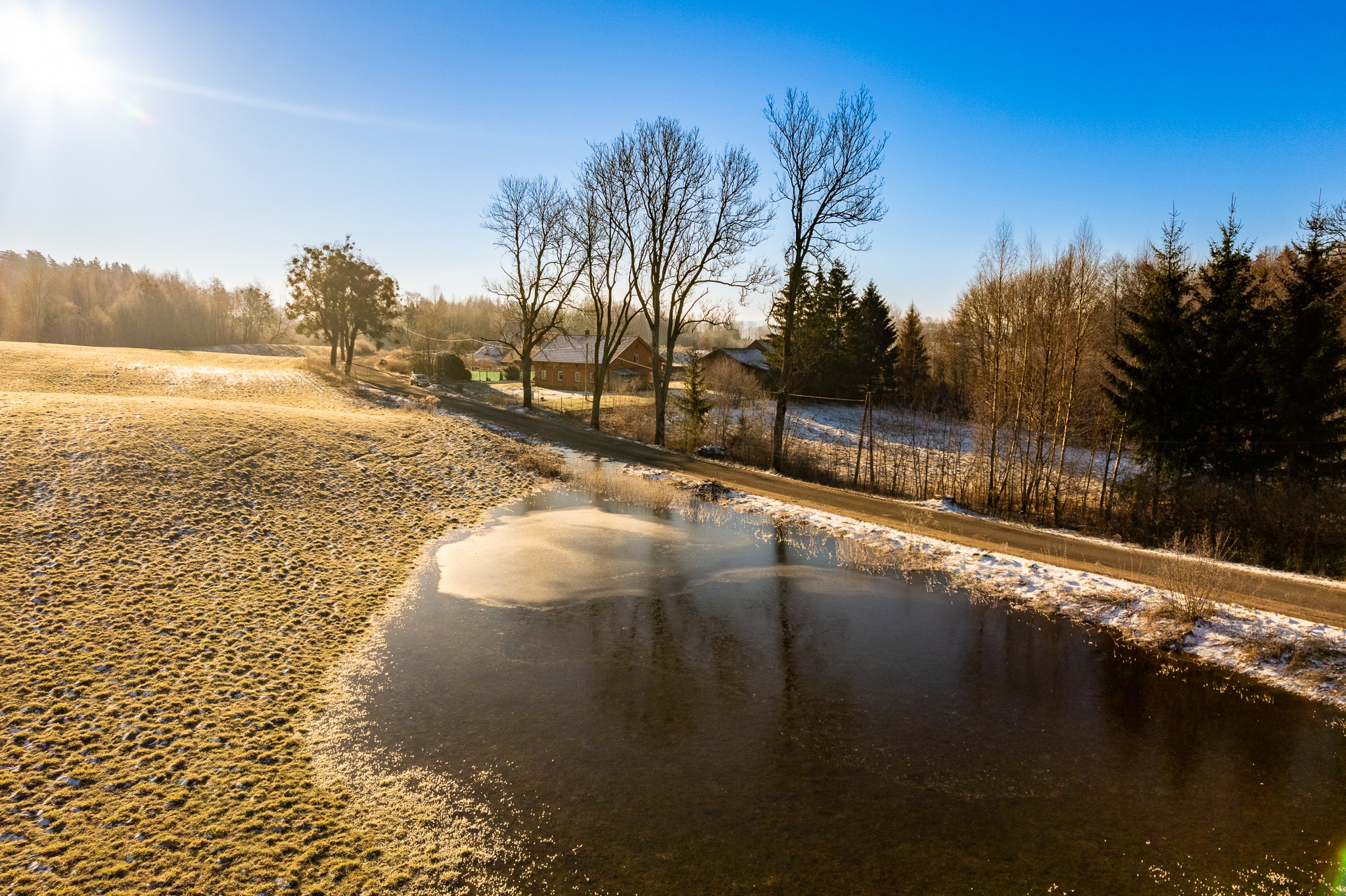 Zimowy krajobraz z częściowo zamarzniętym stawem, polem pokrytym szronem i domem w oddali, sfotografowany w słoneczny poranek.