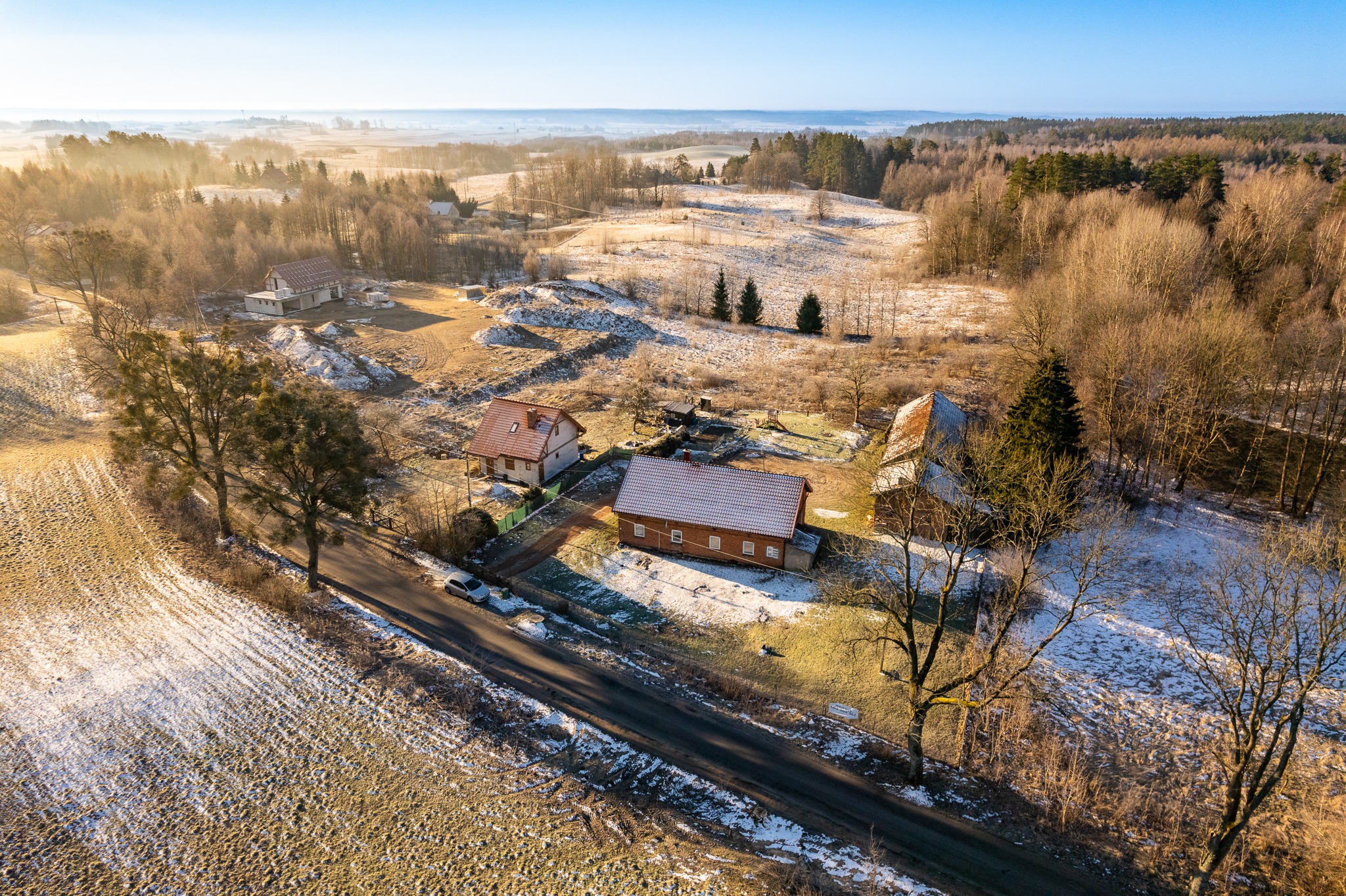 Rozległy krajobraz wiejski z perspektywy drona, ukazujący kilka domów z czerwonymi dachami, pokryte szronem pola i lasy w porannym słońcu.