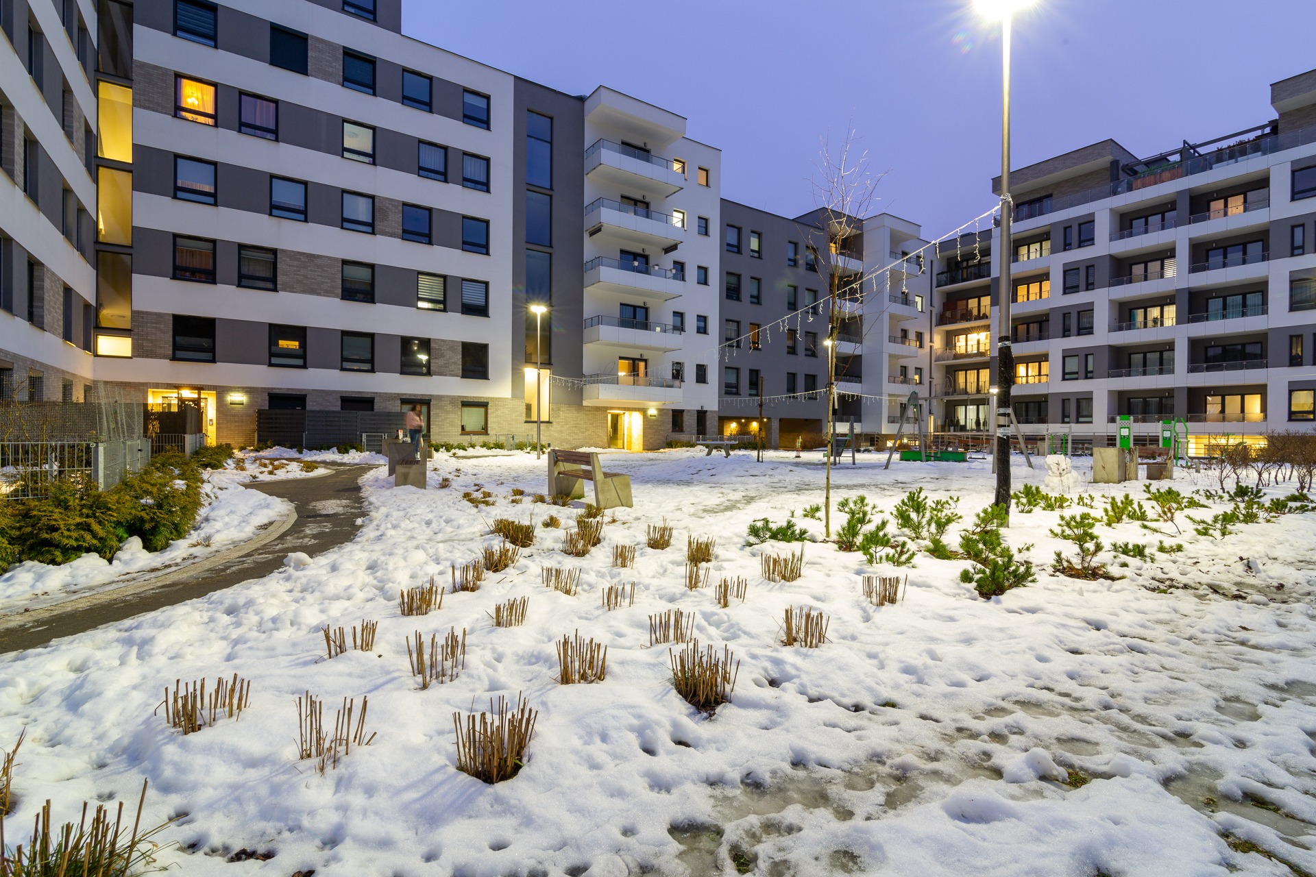 Nowoczesny budynek mieszkalny z balkonami oświetlony wieczornym światłem, otoczony śniegiem i niską roślinnością.