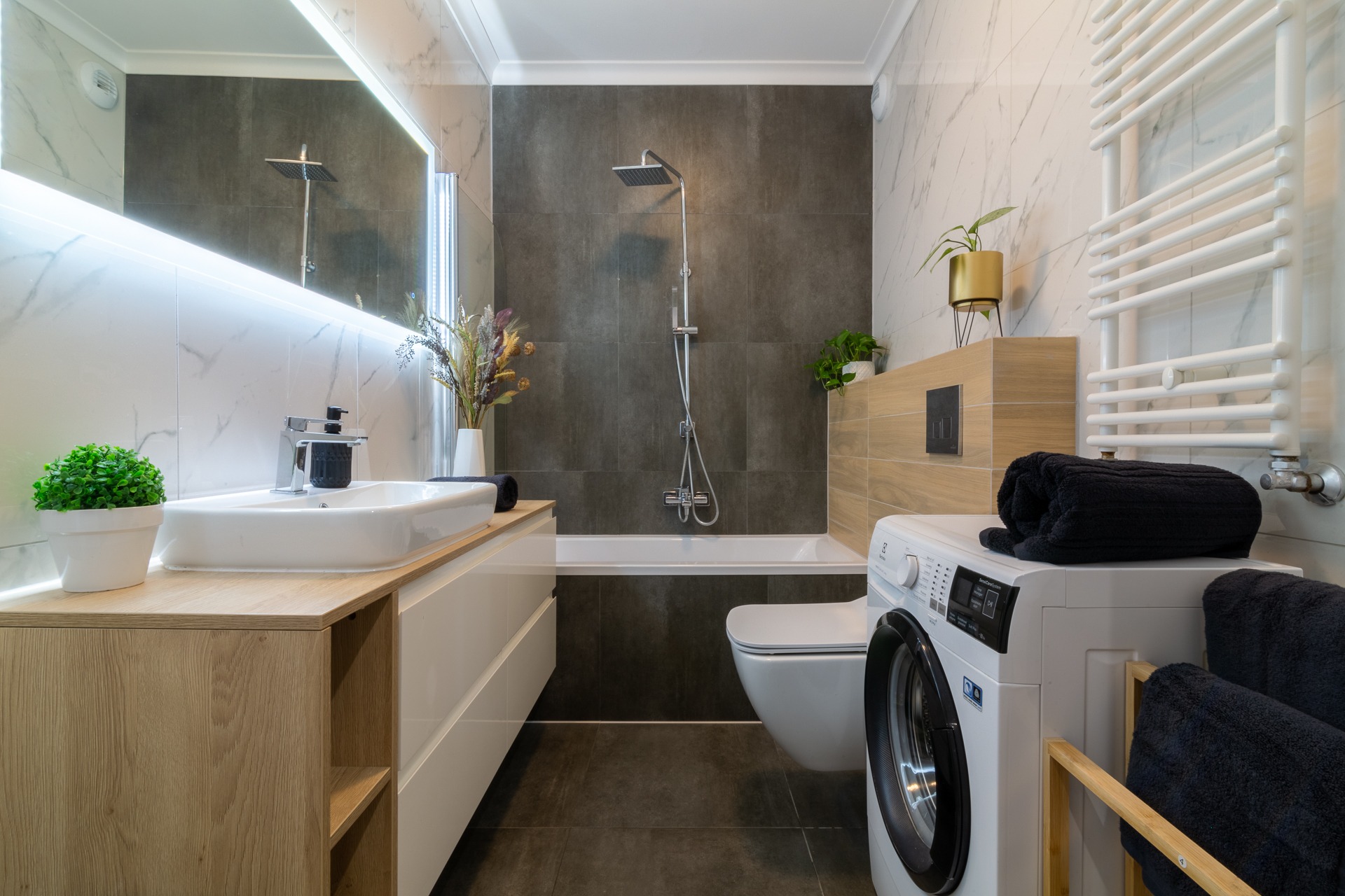 Nowoczesna łazienka z marmurowymi płytkami, drewnianą szafką pod umywalką, wanną z szarą obudową, toaletą, pralką i grzejnikiem drabinkowym, ozdobiona roślinami doniczkowymi i bukietem suszonych...