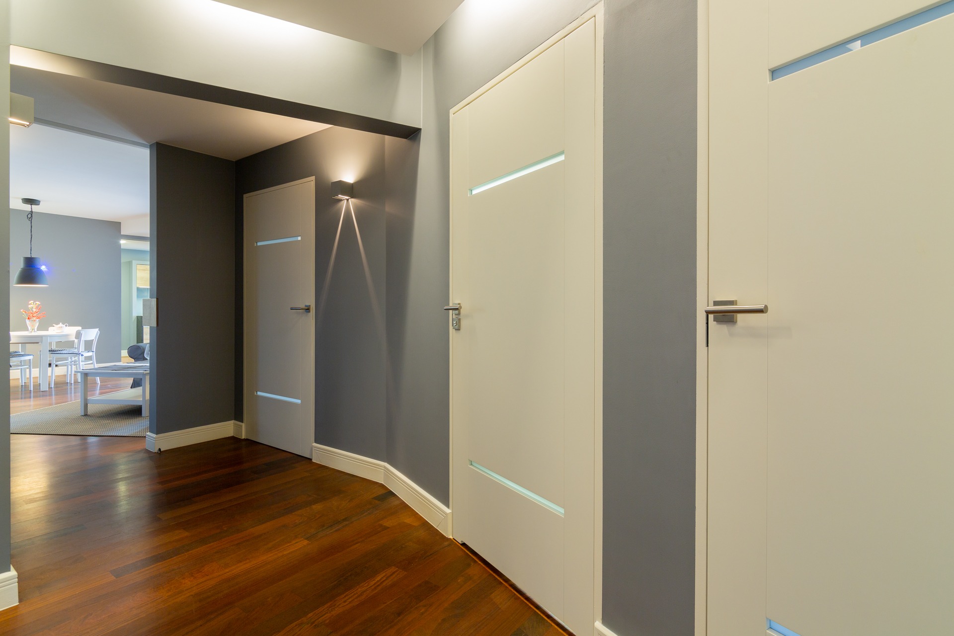 Nowoczesne wnętrze: korytarz z trzema białymi drzwiami z poziomymi przeszkleniami, ciemna podłoga, szare ściany i widok na jadalnię w tle.