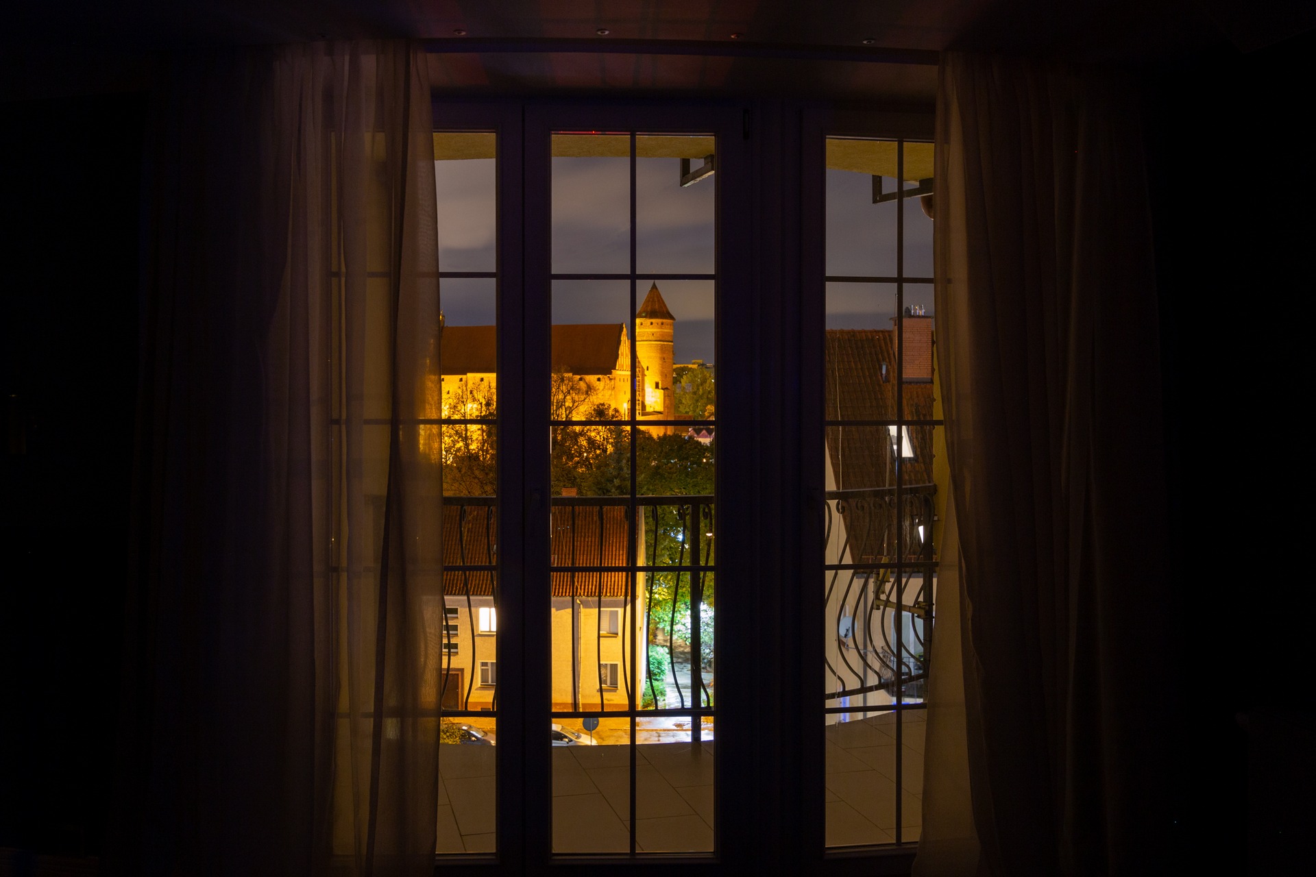 Nocny widok na oświetlony zamek przez okno z białymi zasłonami, widoczny balkon z metalową balustradą.