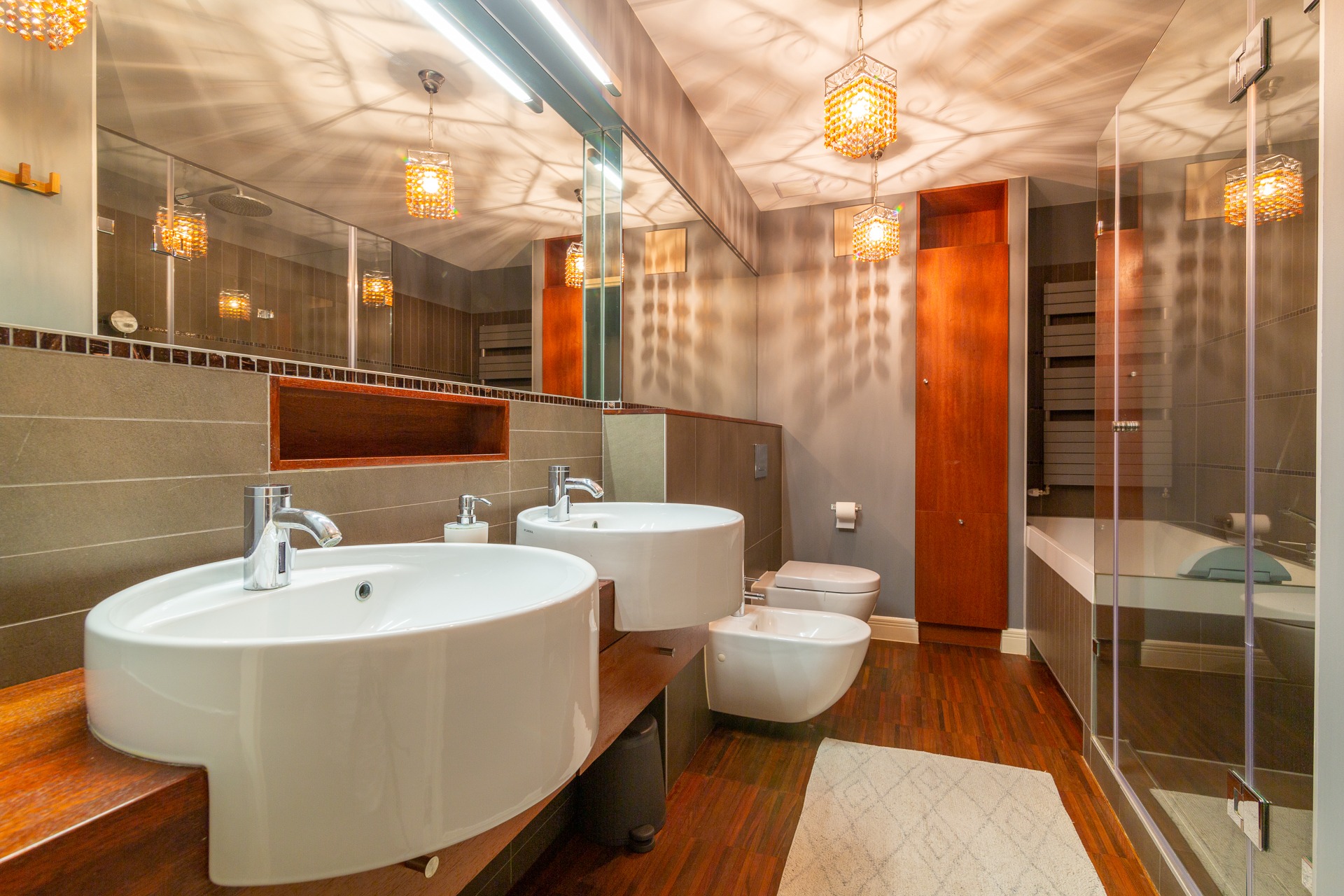 Nowoczesna łazienka z dwoma umywalkami nablatowymi, drewnianym blatem, wiszącą toaletą i bidetem, kabiną prysznicową ze szklanymi drzwiami oraz dekoracyjnymi lampami sufitowymi z odbiciami w lustrze.